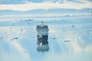 The Antarctic Explorer cruise ship gracefully navigates through icy waters, surrounded by small icebergs. Snow-covered mountains loom majestically in the backdrop, their reflections shimmering on the calm, clear water, creating a scene of breathtaking tranquility.