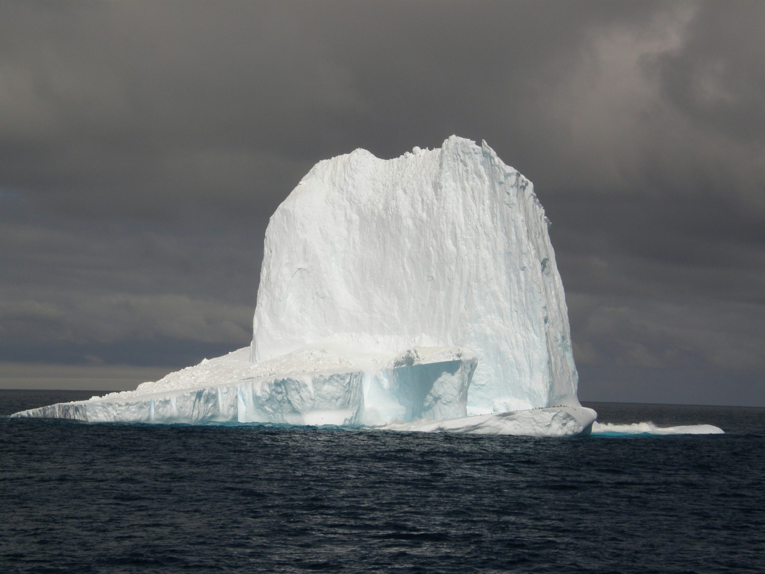 A large iceberg with a towering, rugged peak floats in the dark, calm ocean waters under a cloudy sky. This mesmerizing scene is a glimpse into the breathtaking landscapes experienced when discovering Antarctica's untouched beauty.