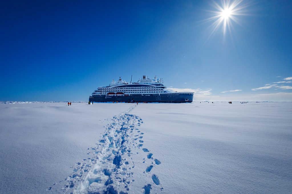 A large cruise ship, renowned among the best Antarctica cruises, is docked on a snowy landscape under a bright blue sky and shining sun. Footprints in the snow lead towards the ship, highlighting the expansive icy surroundings.