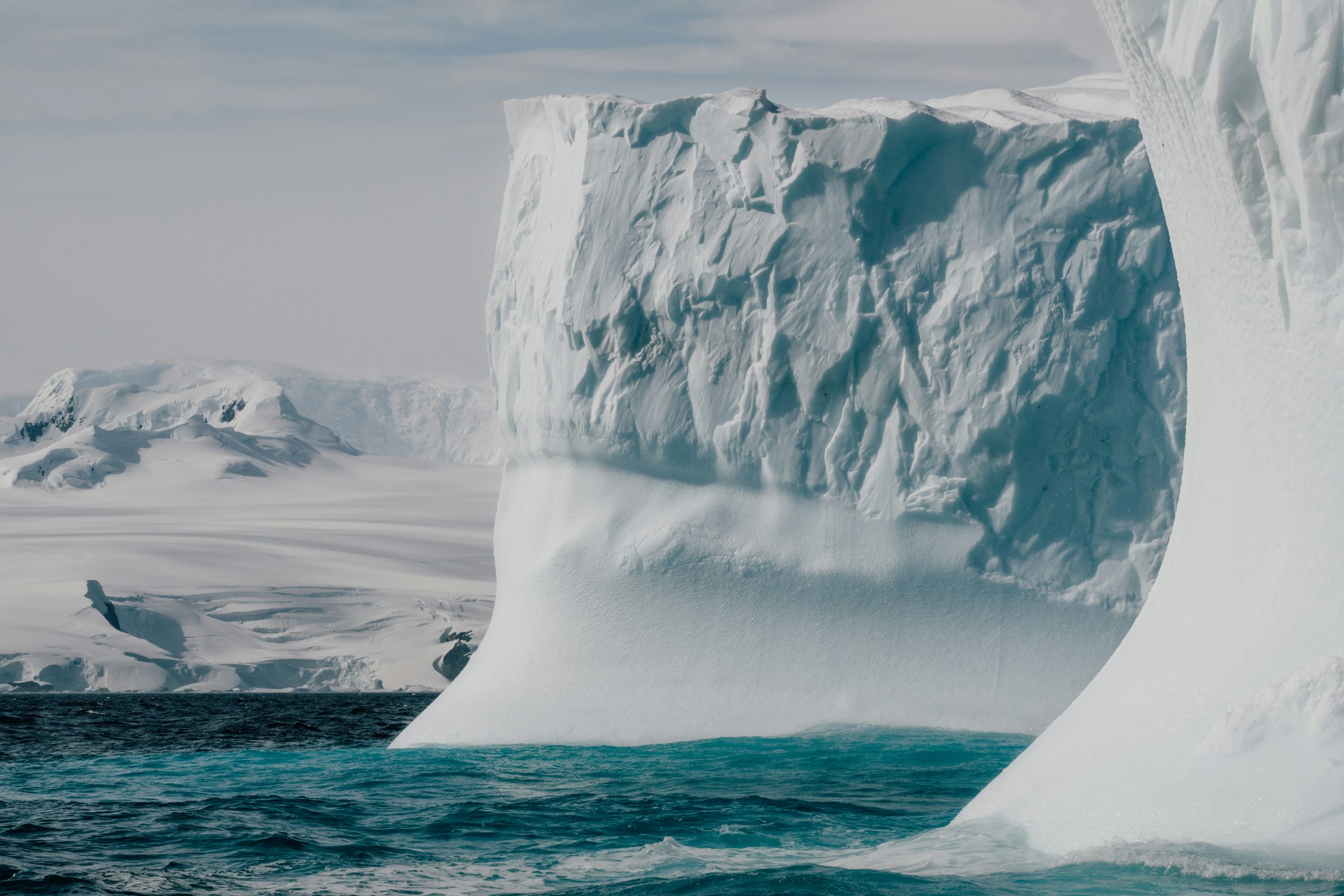 A massive iceberg with jagged, icy surfaces rises from the deep blue sea, offering a breathtaking view perfect for Luxury Antarctica Cruises. Snow-covered mountains extend across the background, creating a stark contrast between the ice and turquoise waters beneath the clear sky.