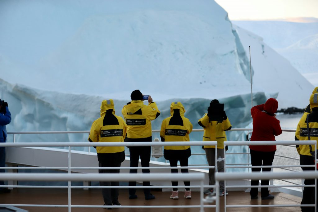 Amid icy, Arctic-like surroundings, people in yellow jackets stand on a ship's deck capturing photos of a towering iceberg. One person in a red jacket holds a long pole. This breathtaking scene could easily be part of the best trips to Antarctica for 2026.