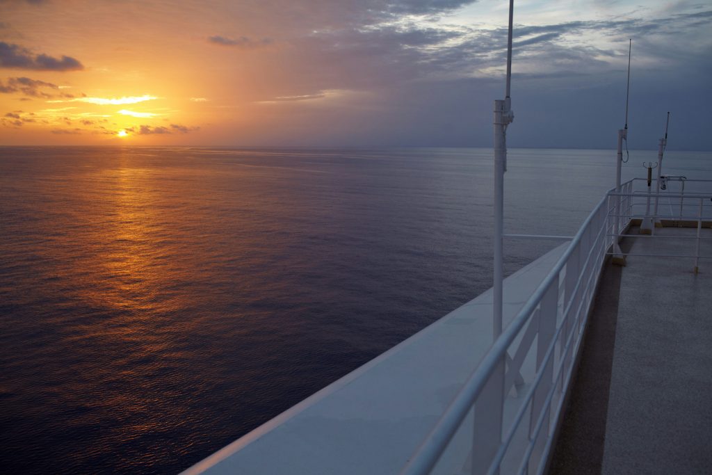 Sunset over a calm ocean, viewed from a ship's deck on one of the best Alaskan cruises. The sky transitions from vibrant orange at the sun-kissed horizon to deeper blues as clouds gather on the right. The ship's railings elegantly line the right side of this serene scene.