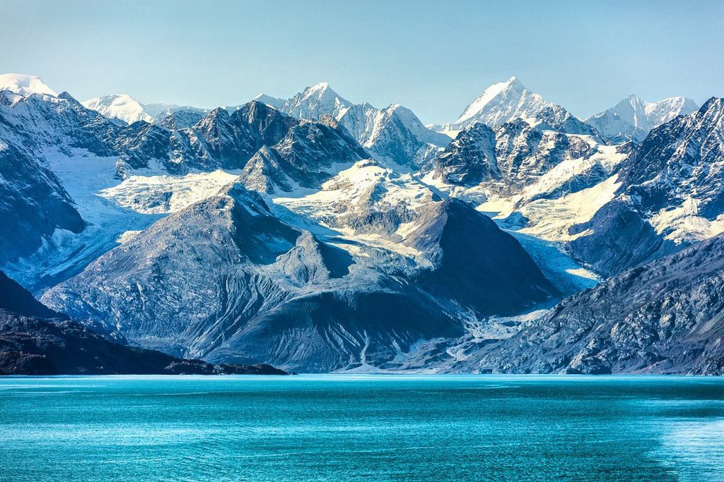 A stunning view of snow-capped mountains under a clear blue sky. Rugged peaks and glaciers slope down to a turquoise sea in the foreground, creating a vivid and serene landscape.
