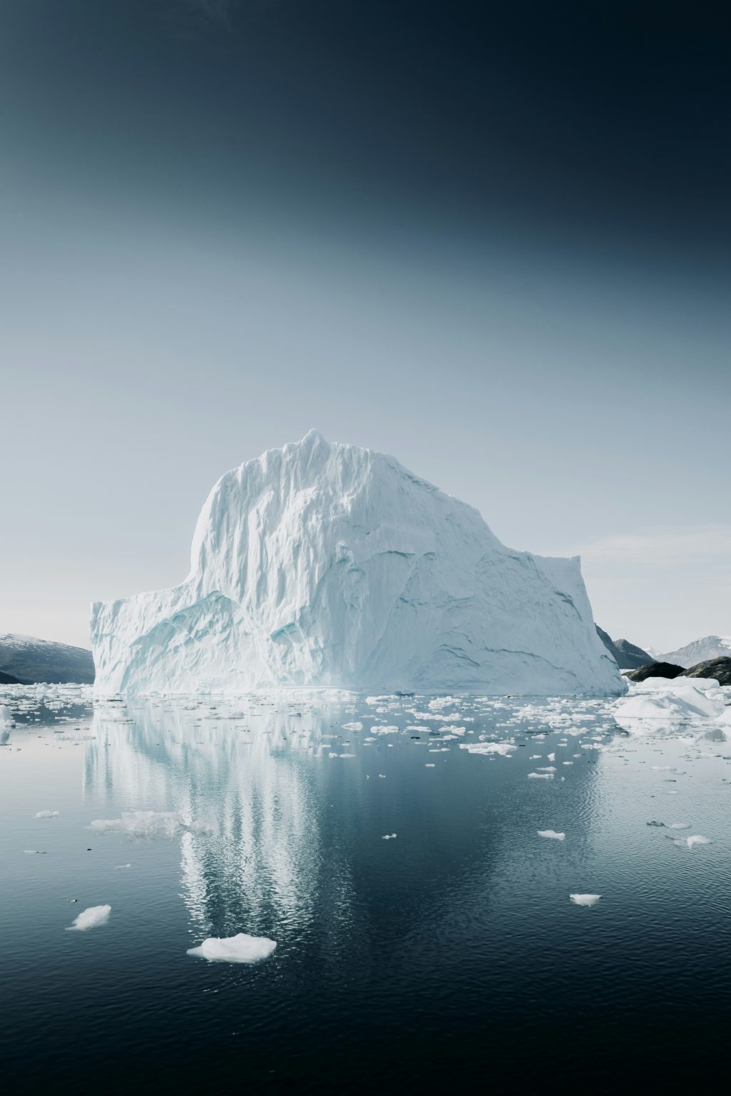 A large iceberg traversing the Northwest Passage floats in calm waters, mirroring its image on the surface. The sky above is clear, with scattered smaller ice chunks surrounding it. Rugged, snowy mountains loom in the distant background.