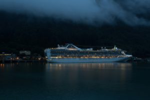 A large, illuminated cruise ship is anchored at a port during the night, reminiscent of the best Antarctic cruises. Surrounding it are dark, forested hills partially covered by low-lying clouds. The water reflects the ship's lights, creating a serene, moody atmosphere. Juneau, Alaska