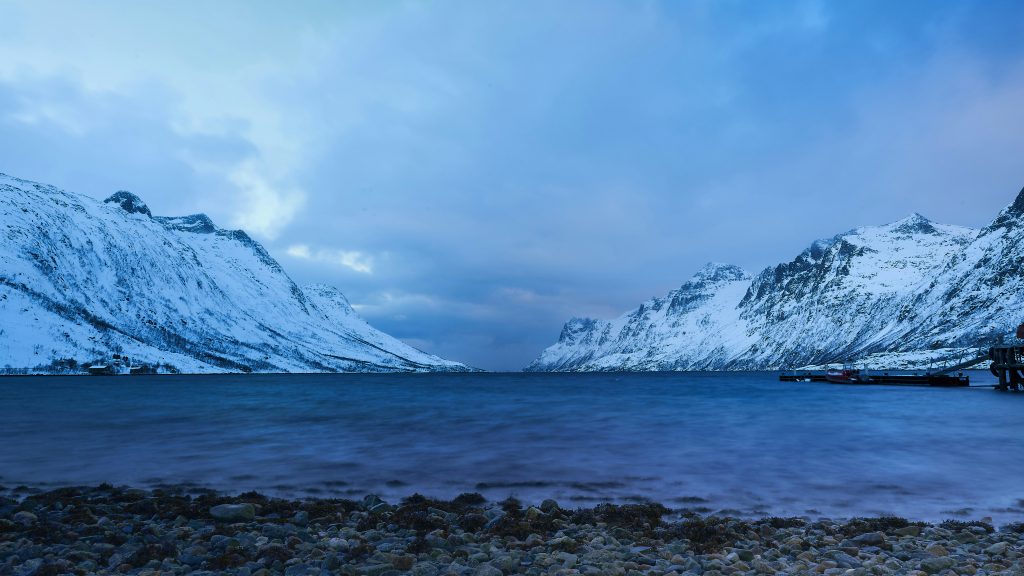 Under a cloudy sky, a serene fjord scene unfolds with snow-covered mountains lining the sides. The calm water reflects the soft blue hues, while a rocky shoreline graces the foreground. On the right, a small pier stretches out—inviting thoughts of embarking on a trip to see the Fjords in Norway.