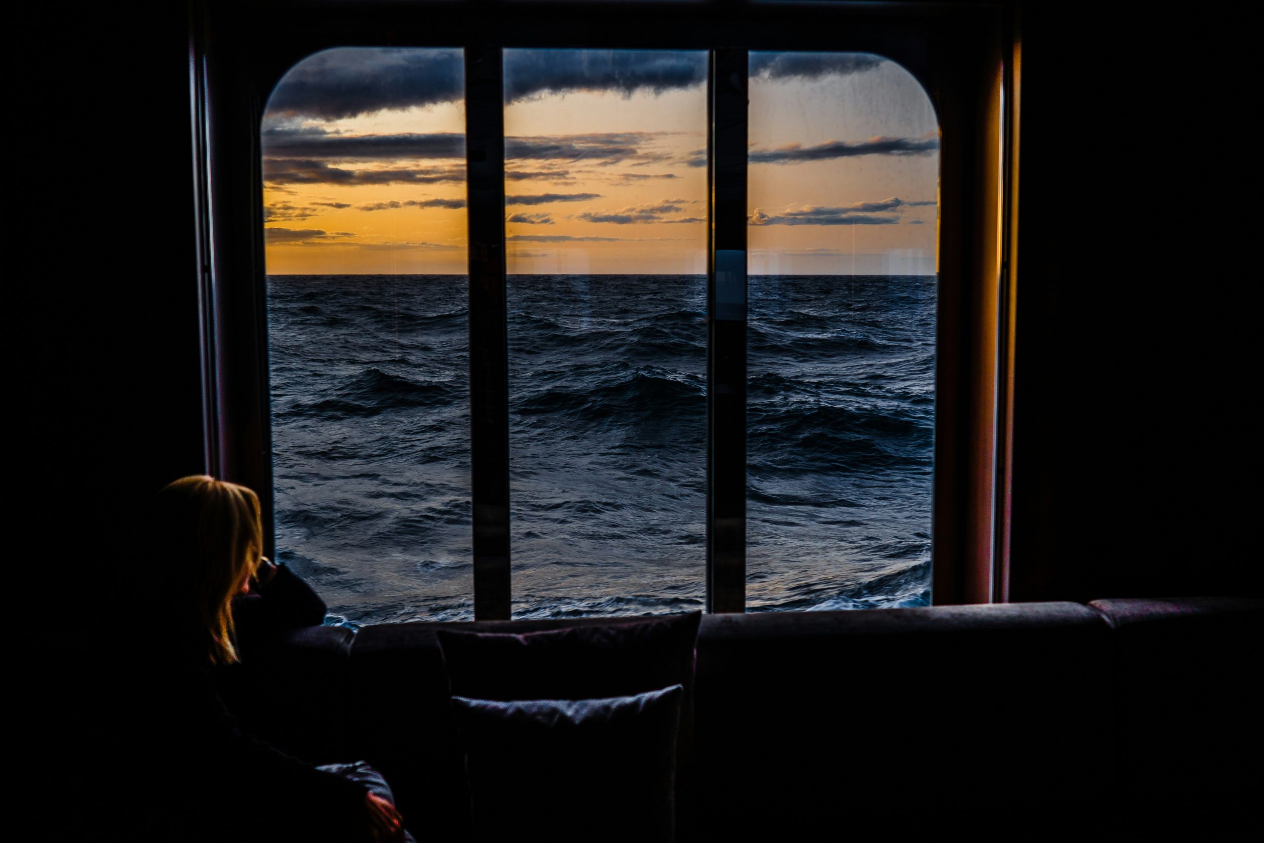 A person sits inside, silhouetted against a large window with an ocean view. As the sun sets, painting the sky in warm tones of orange and yellow, they dream of future adventures and wonder about the best Antarctica cruises for 2025 while watching waves ripple across the deep blue sea.