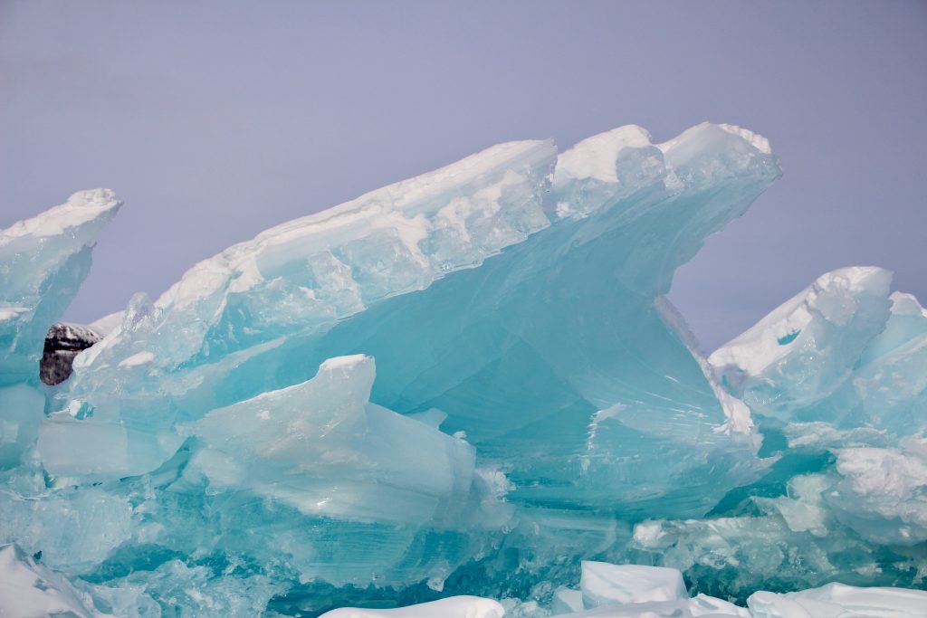 Large, angular slabs of turquoise ice are stacked upon one another against a pale blue sky, like the stunning sights you'd see on an Alaska cruise. The ice features jagged edges and varied transparency, creating an abstract and dynamic composition with snow-dusted peaks.