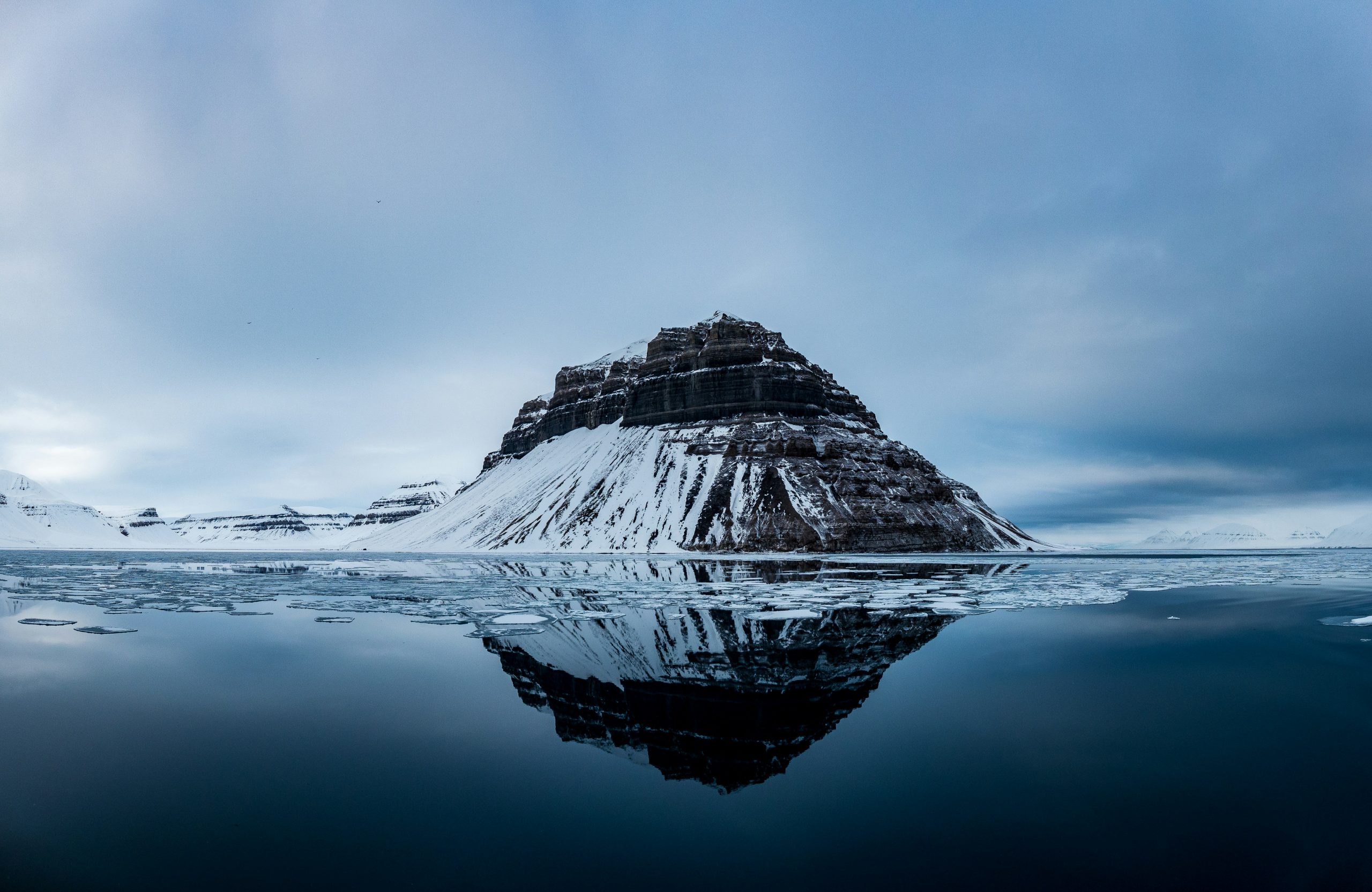 Melting sea ice at the entrance to Billefjorden. A serene, remote landscape unfolds as a mountain with snow-covered slopes reflects in a calm body of water under a cloudy sky, reminiscent of vistas found on Alaska cruise deals. Icy patches drift across the surface, capturing the essence of cold tranquility.