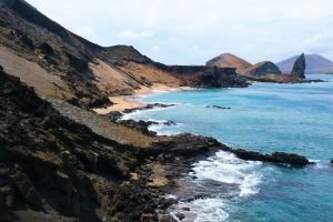 A coastal landscape with rugged, dark cliffs and a sandy beach. The turquoise ocean waves gently lap against the rocks, reminiscent of vistas seen on Galapgos Islands.