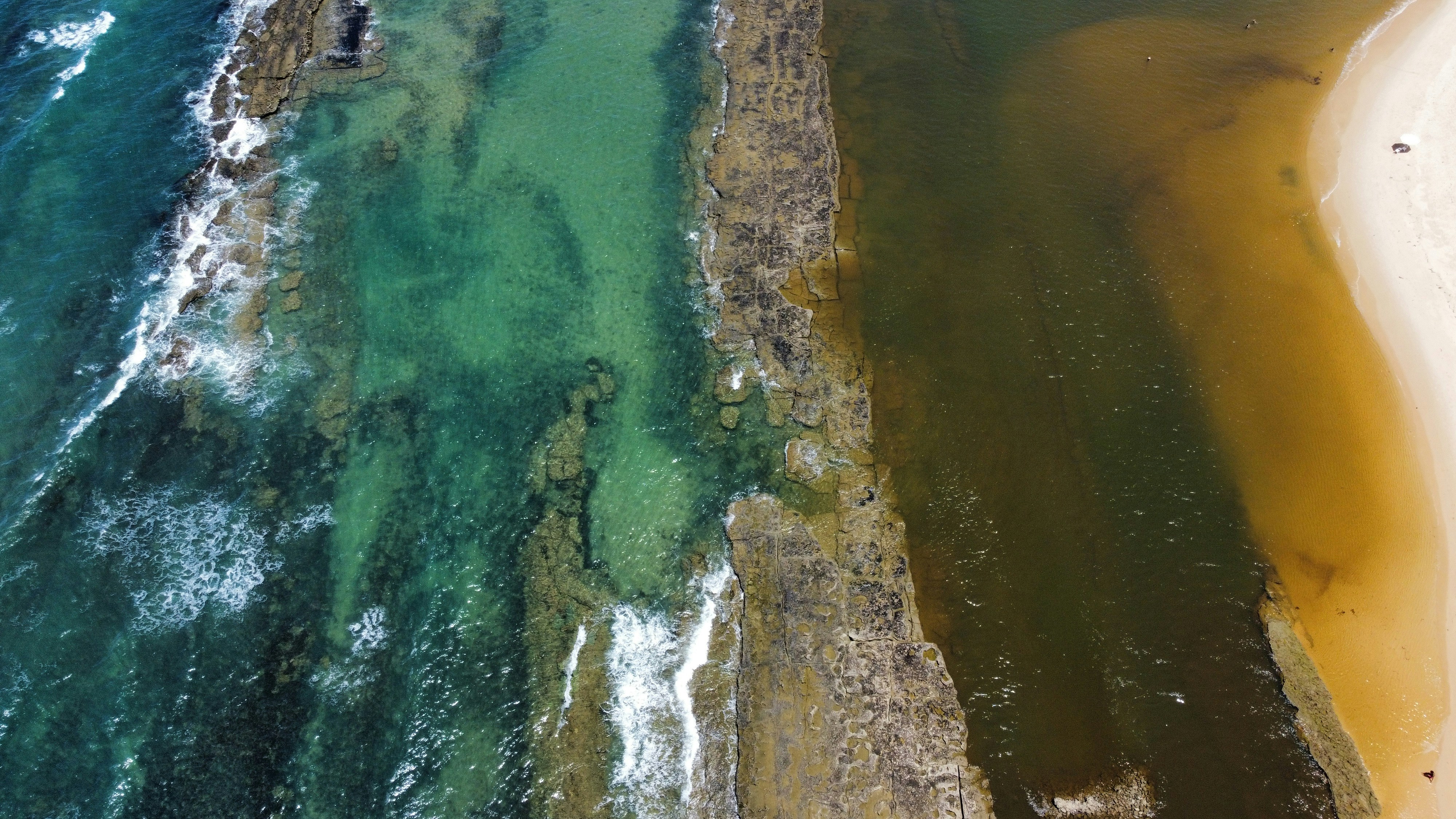 Aerial view of a beach with clear turquoise water meeting darker water and sand. Rocky formations run through the water in a diagonal line, separating the two shades. The sand is light, and small waves approach the shore.