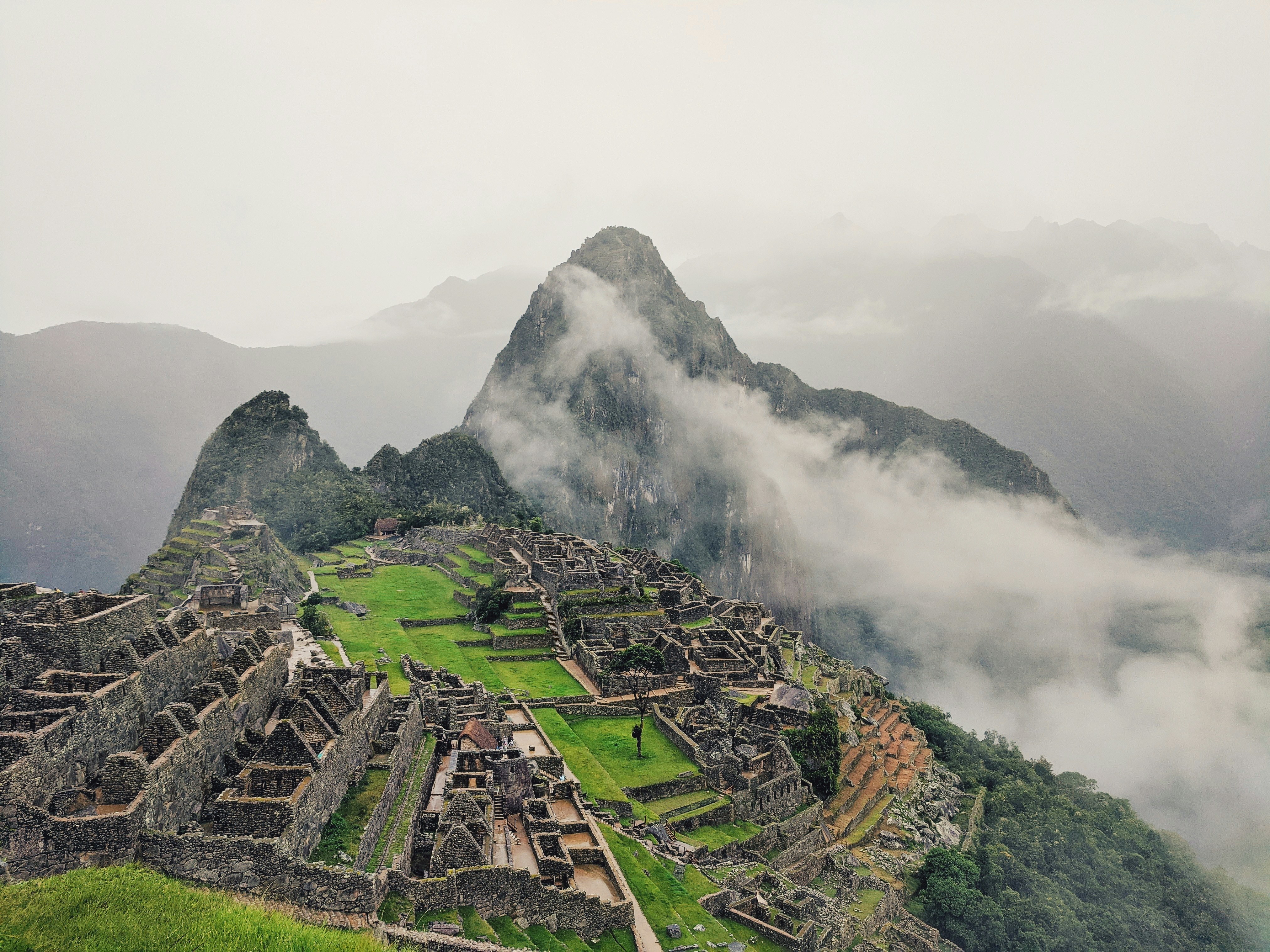 A misty view of Machu Picchu reveals ancient stone structures and terraces nestled in lush greenery, reminiscent of The Meeting of Waters. The iconic Huayna Picchu mountain looms in the background, partially enveloped by clouds, enhancing the mystical atmosphere.