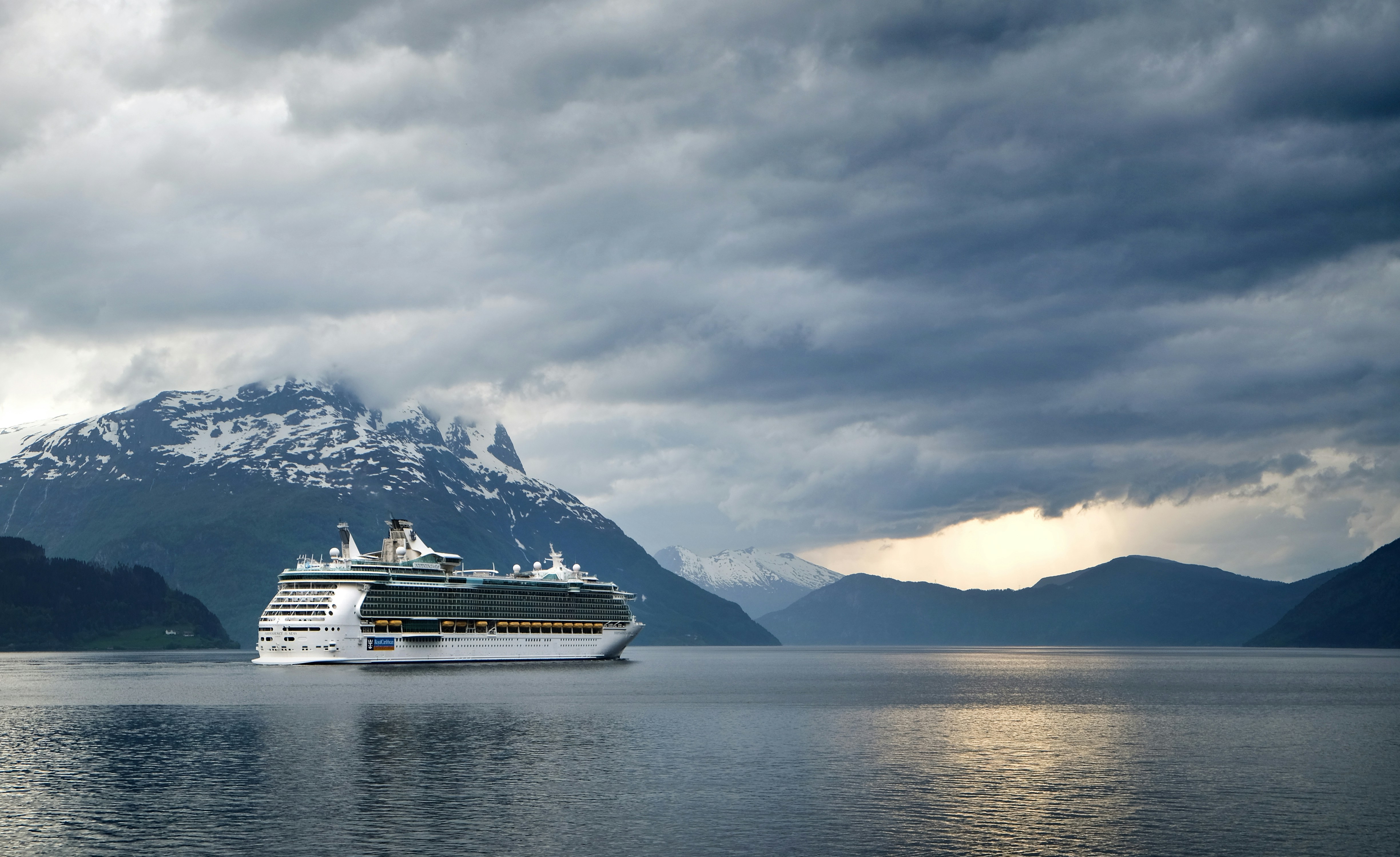 A large cruise ship sails through a calm fjord surrounded by scenic mountains partially covered with snow. Reminiscent of trips to the Amazon, the dramatic overcast sky casts soft reflections on the water, offering travelers an unforgettable journey through serene landscapes.