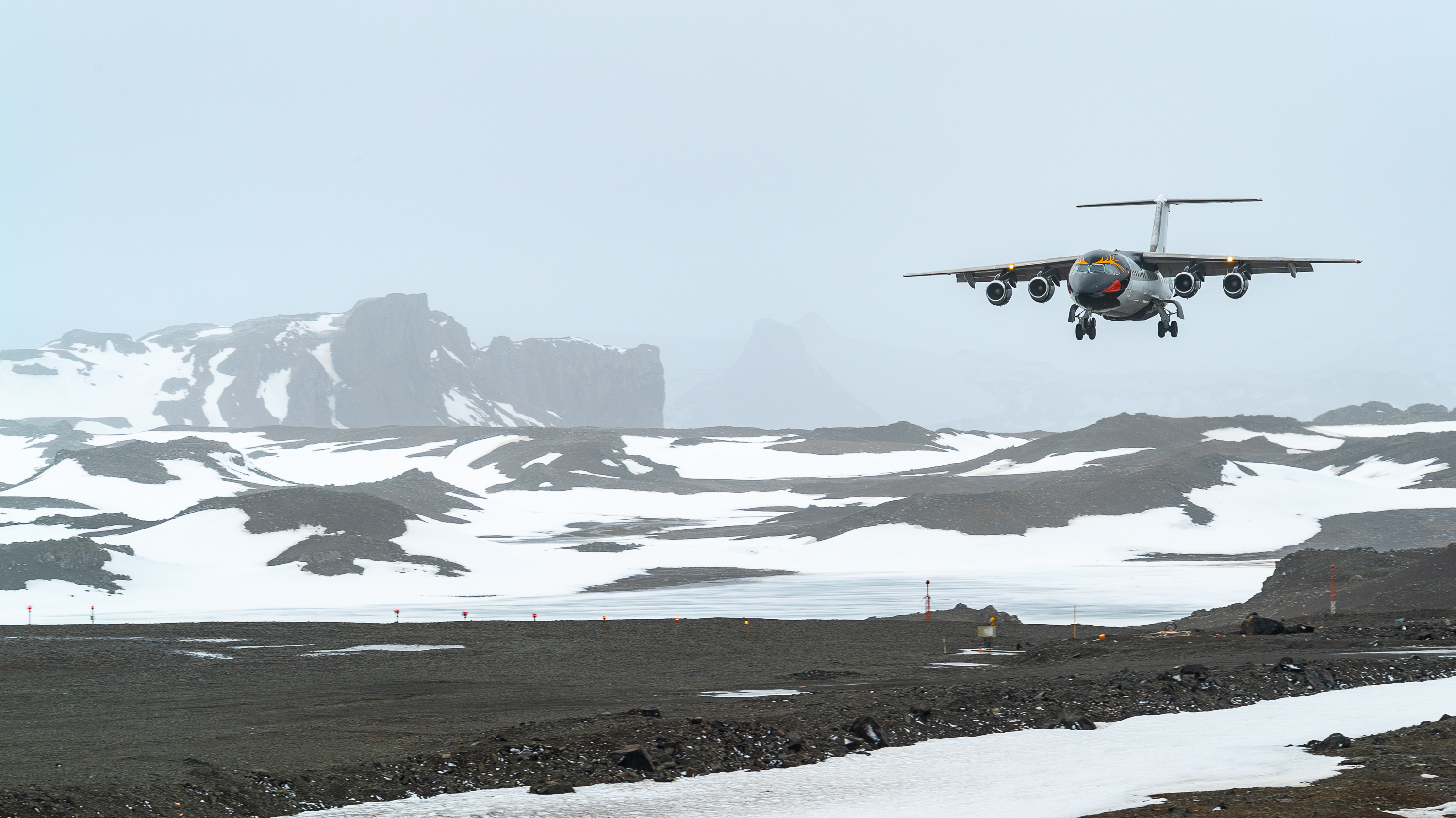 A gray aircraft is landing on a snowy, rocky landscape in Antarctica, with mountains in the background under a cloudy sky. Patches of snow and ice cover the ground.