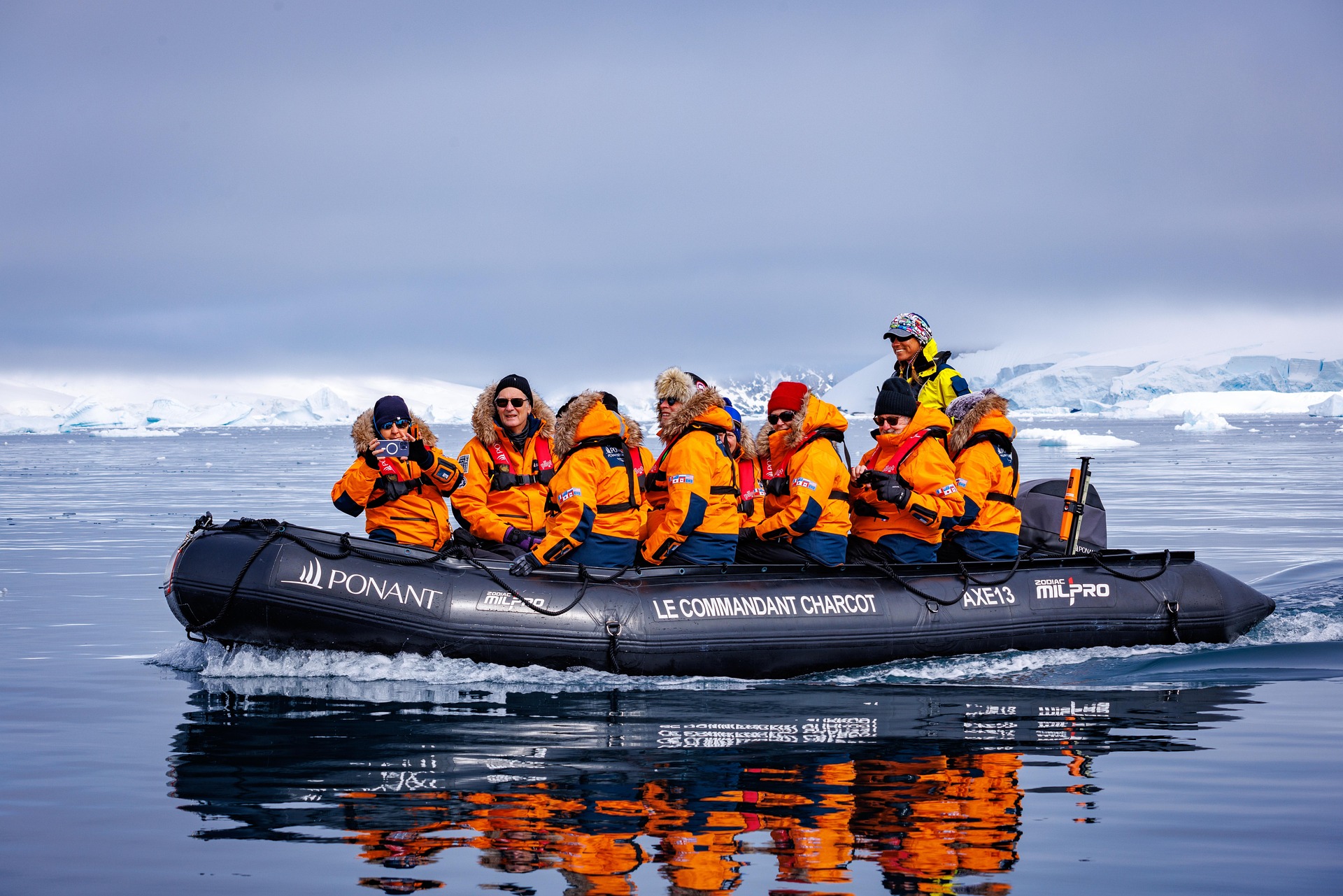 A group of people wearing orange coats and life jackets ride an inflatable boat labeled "Le Commandant Charcot" across icy blue water during their Antarctica Air-Cruise, with snowy, glacier-covered land visible in the background.