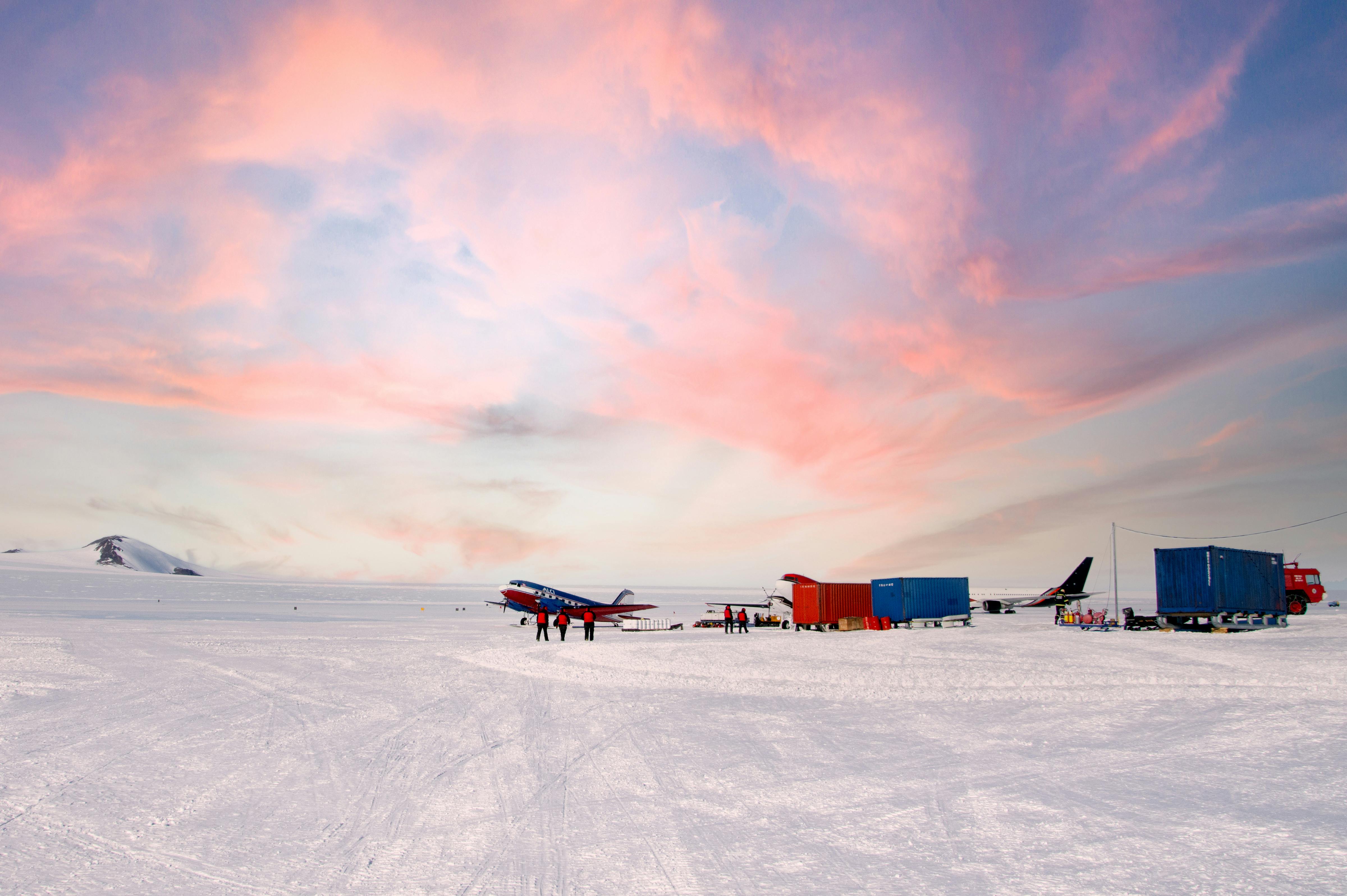 An airplane and colorful cargo containers sit on snowy ground under a dramatic pink and orange sunset in the Falklands, South Georgia, or Antarctica. People stand by the plane, with a distant mountain visible on the left.