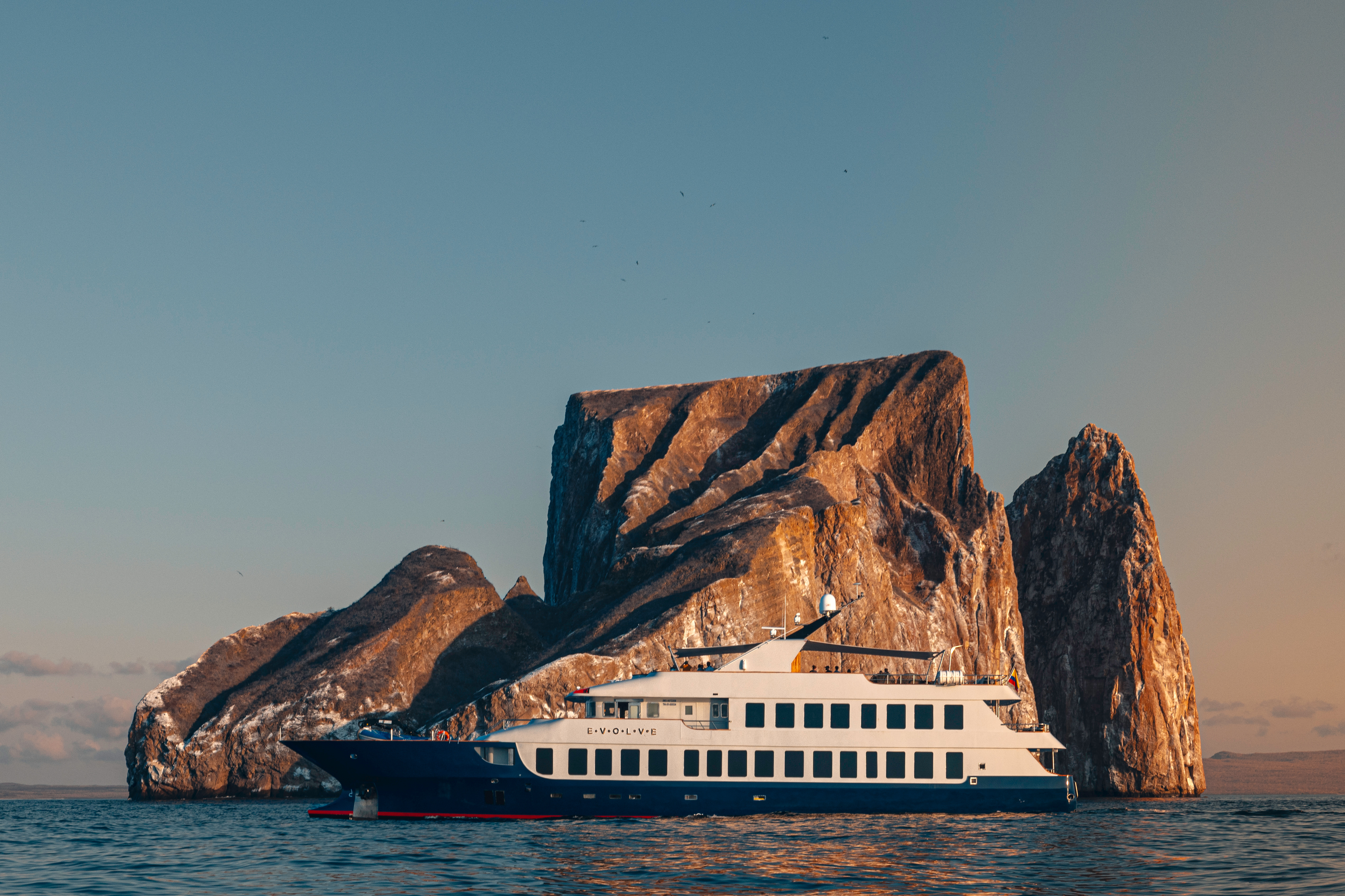 Ecoventura's Evolve - a large yacht sails on calm water near a dramatic, rocky island with steep cliffs, under a clear sky at sunset.