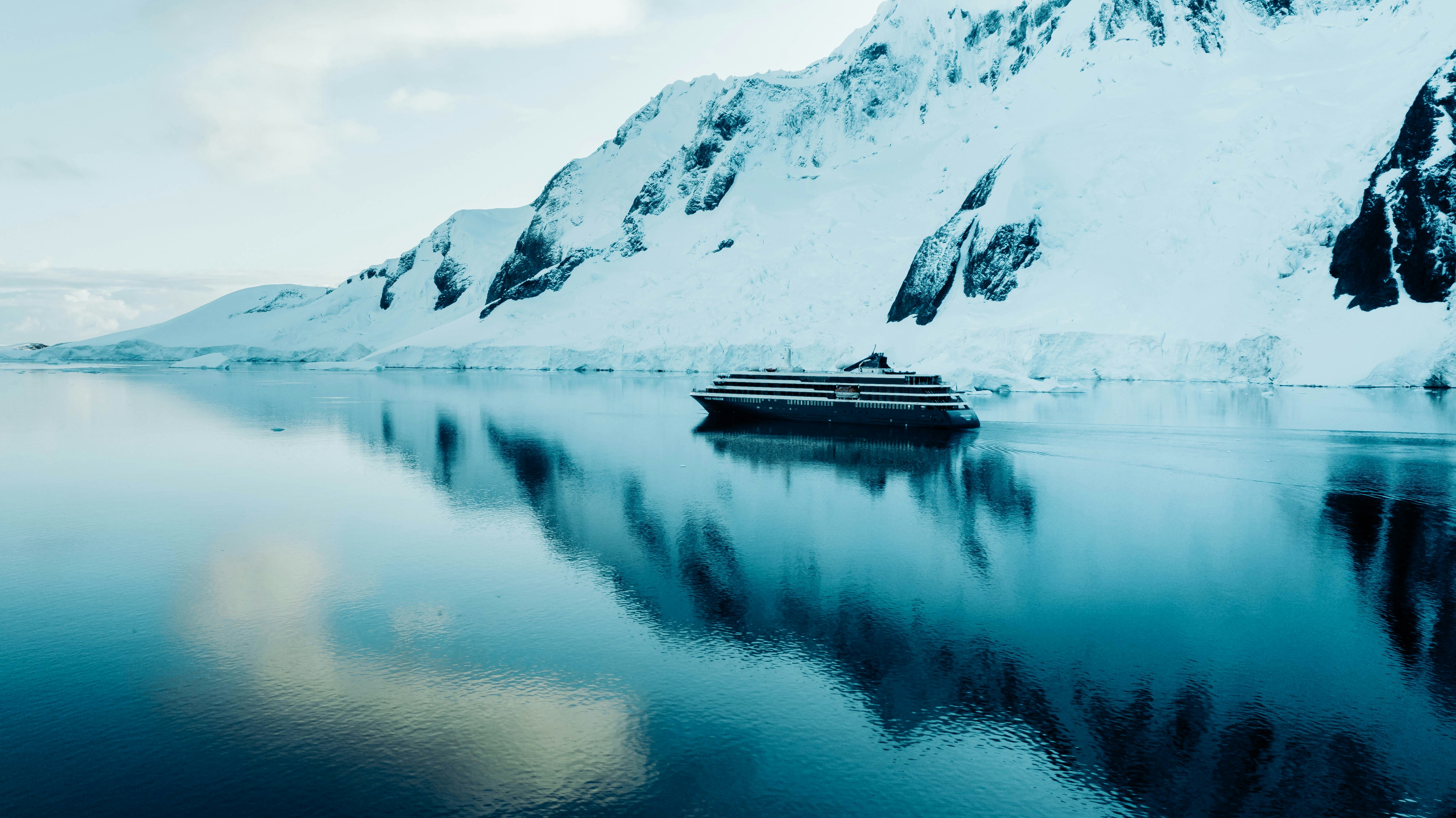 A cruise ship sails on calm, icy blue water in Antarctica 2026, surrounded by snow-covered mountains and glaciers, with the breathtaking landscape reflected in the water under a cloudy sky.