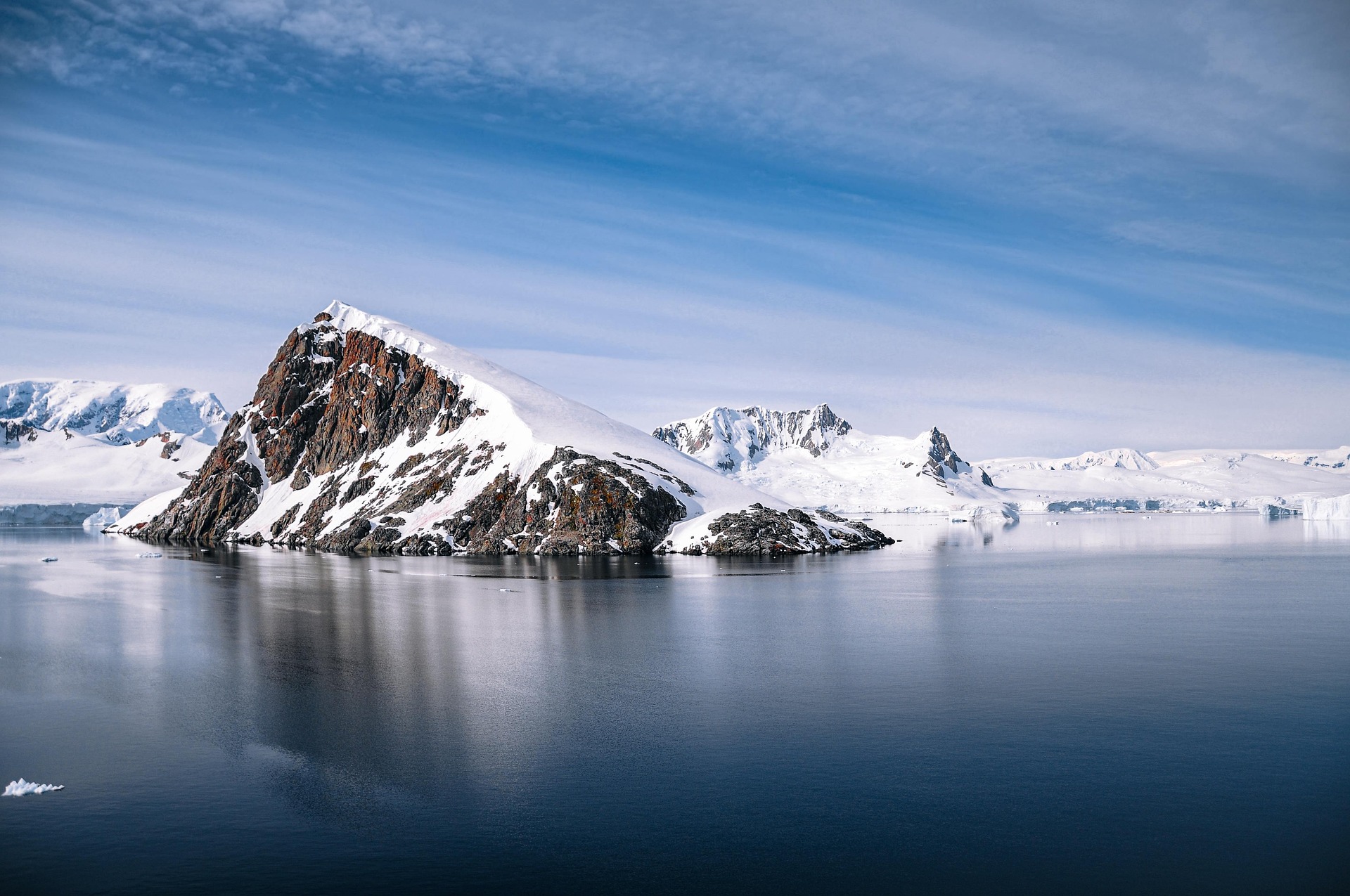 A rocky, snow-covered island sits in calm, icy blue water, with distant snow-capped mountains—an awe-inspiring scene often glimpsed by those curious about the cost of an Antarctica cruise to this cold, remote landscape.