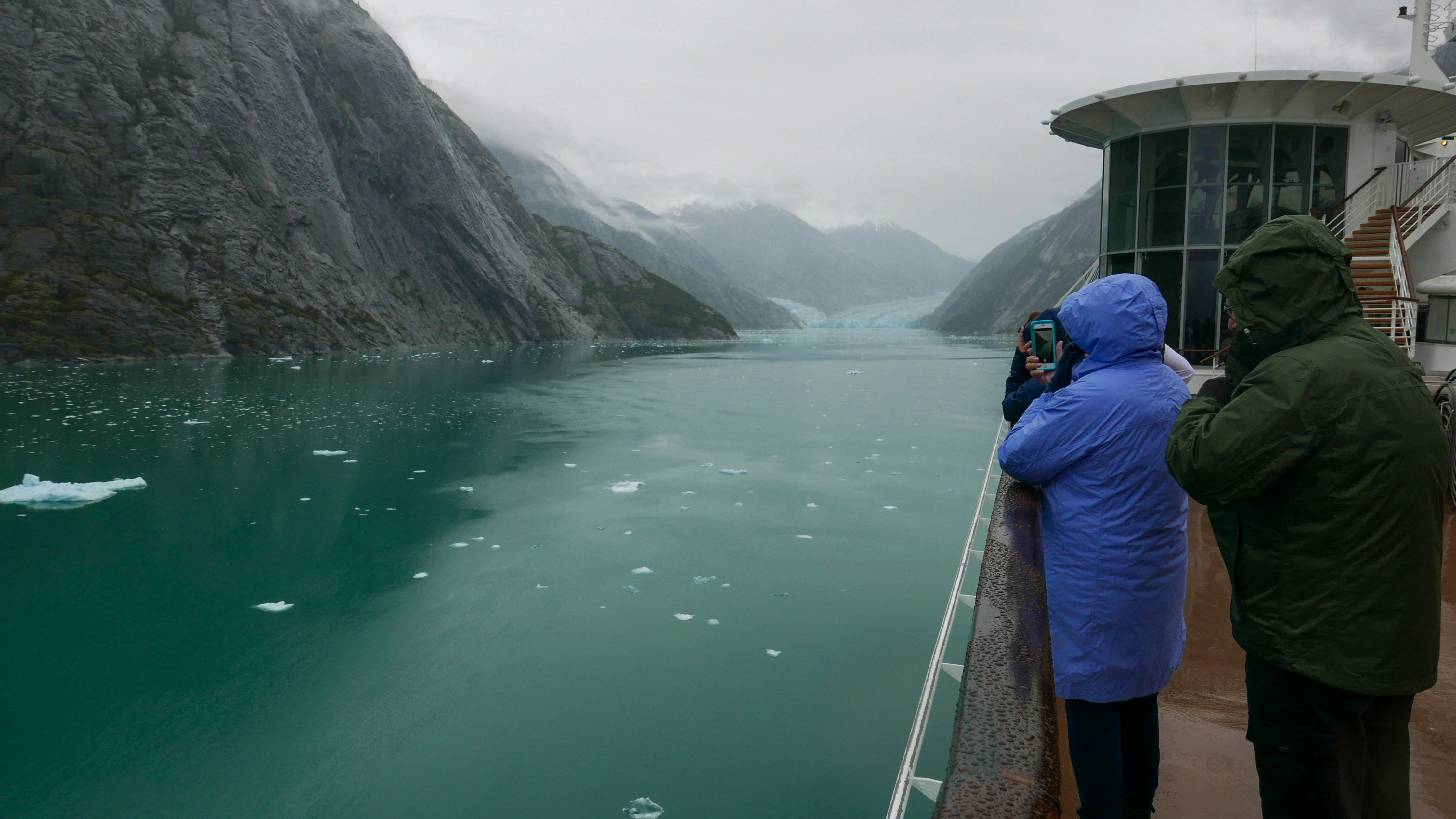 People in raincoats stand on a ship's deck, gazing at a misty, glacier-filled fjord with snowy mountains and floating ice—an awe-inspiring scene often raised in Arctic vs Antarctic travel debates.