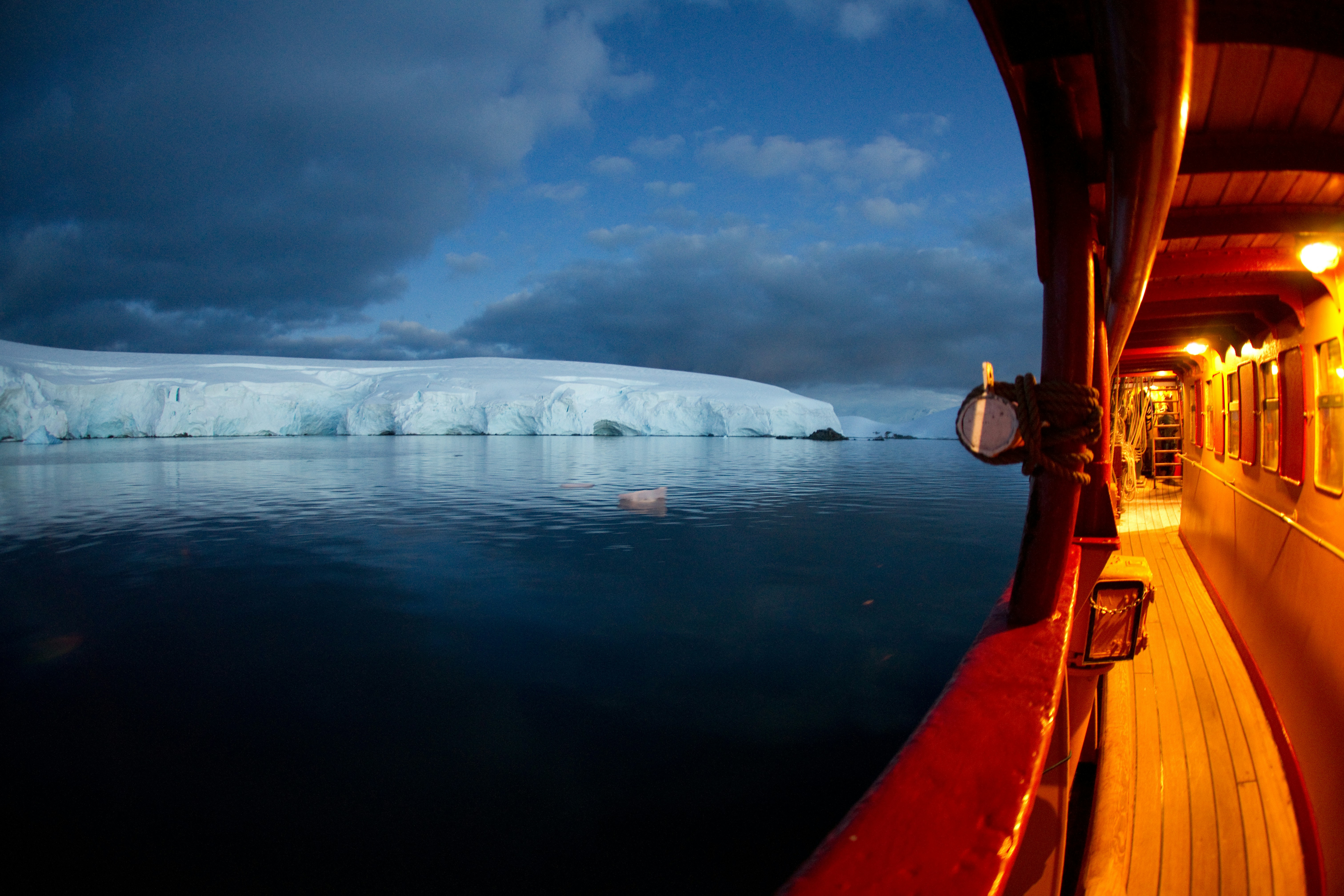 A wooden boat with warm lights reflecting off its deck sails near a large ice-covered landmass under a cloudy, blue evening sky. The calm, dark water mirrors the ice and sky.