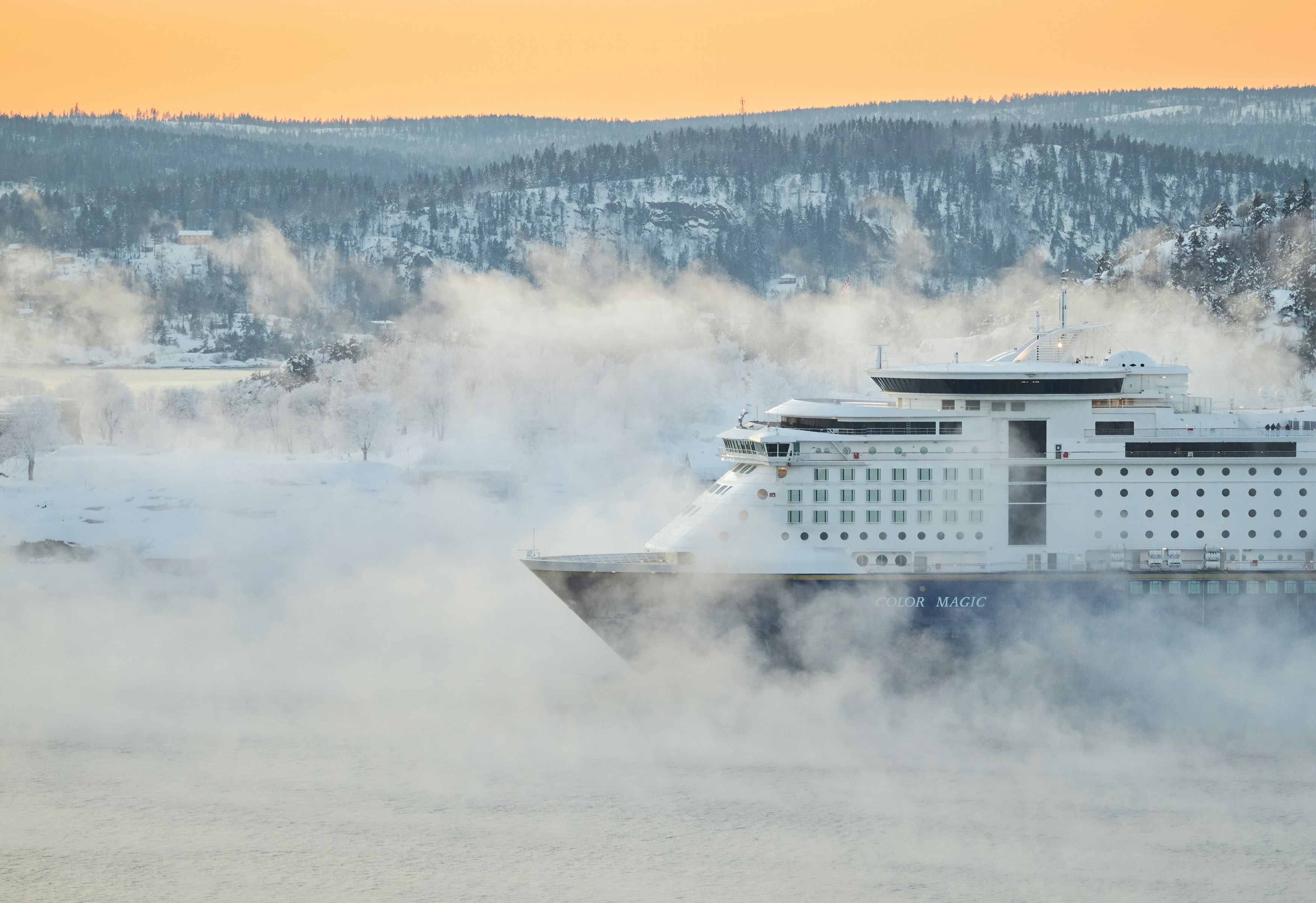 A large cruise ship sails through misty, icy water near a snowy, forested shoreline at sunrise or sunset, with a soft orange sky in the background—perfect for those arriving on a flight to Antarctica seeking breathtaking vistas.