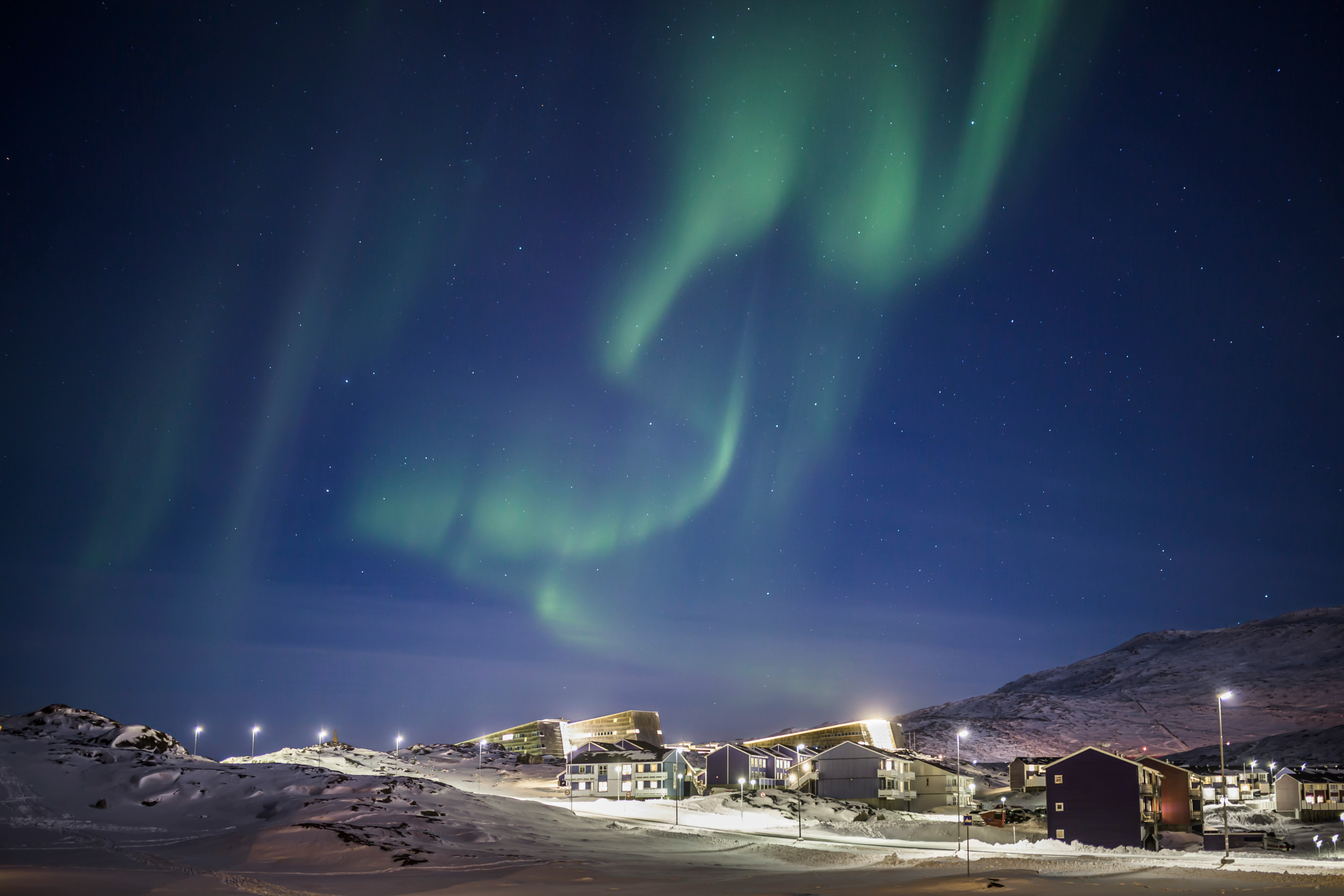 Green northern lights glow in a starry night sky above a snowy village with illuminated houses and streetlights, surrounded by snow-covered hills—a magical scene often witnessed during Alaska Polar Bear Tours.