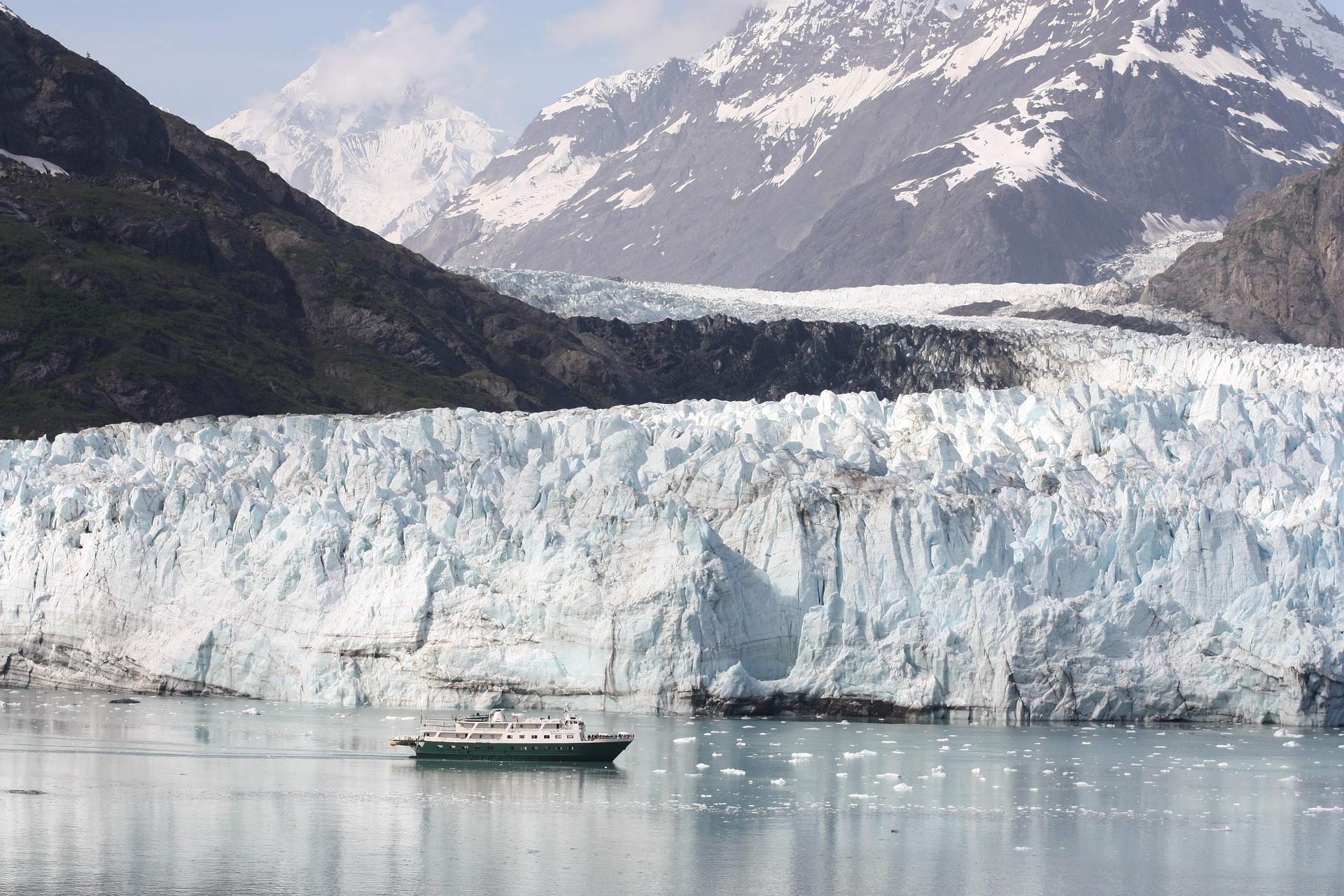 A boat floats on calm water before a massive glacier, with snow-capped mountains rising in the background—scenery reminiscent of Svalbard vs. Greenland vs. the Canadian Arctic under a partly cloudy sky.