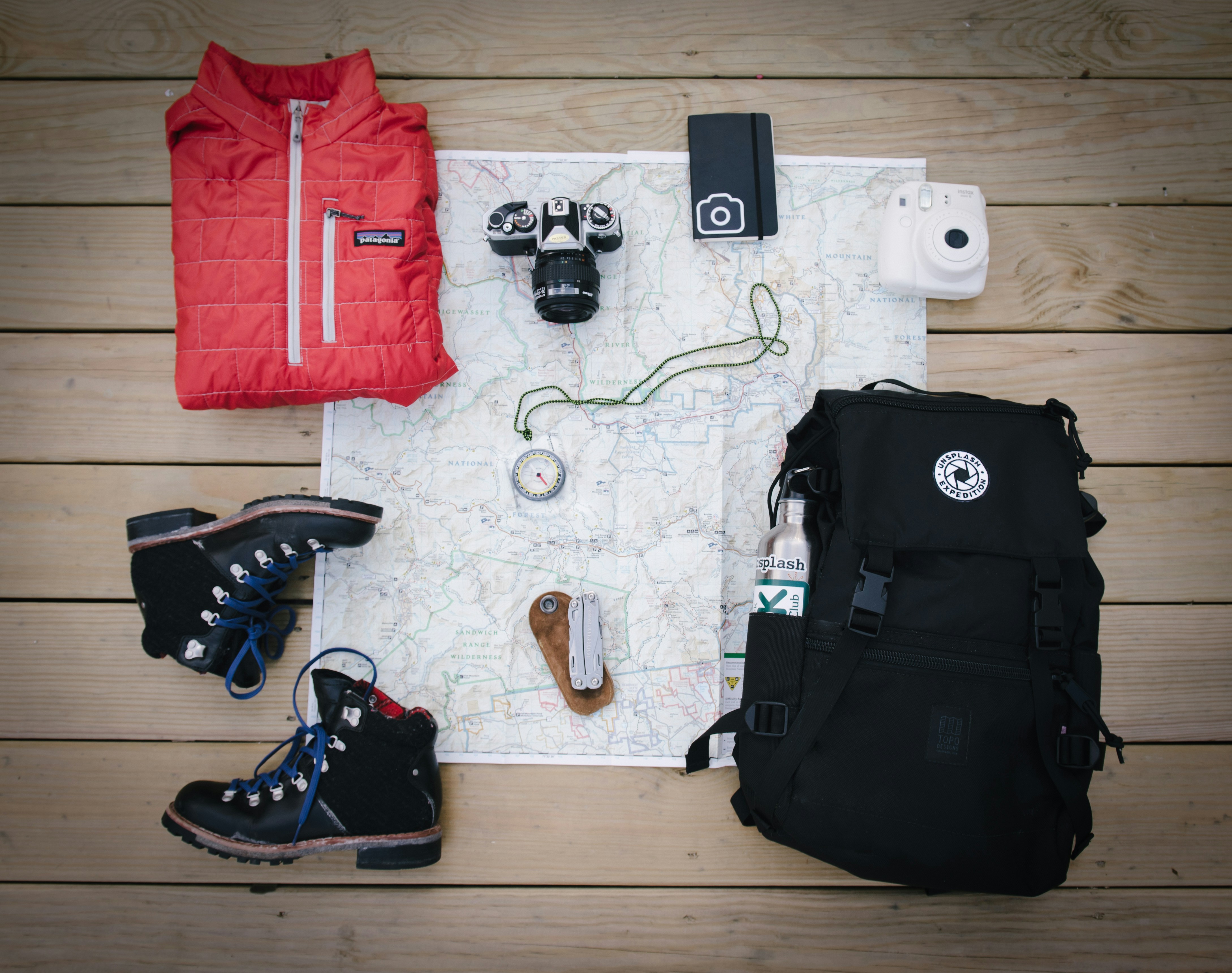 Flat lay of travel gear on wooden boards: red jacket, two cameras, smartphone, instant camera, compass, folding knife, hiking boots, and black backpack with water bottle—essentials for Solar Eclipse 2026 Cruises—arranged around a map.