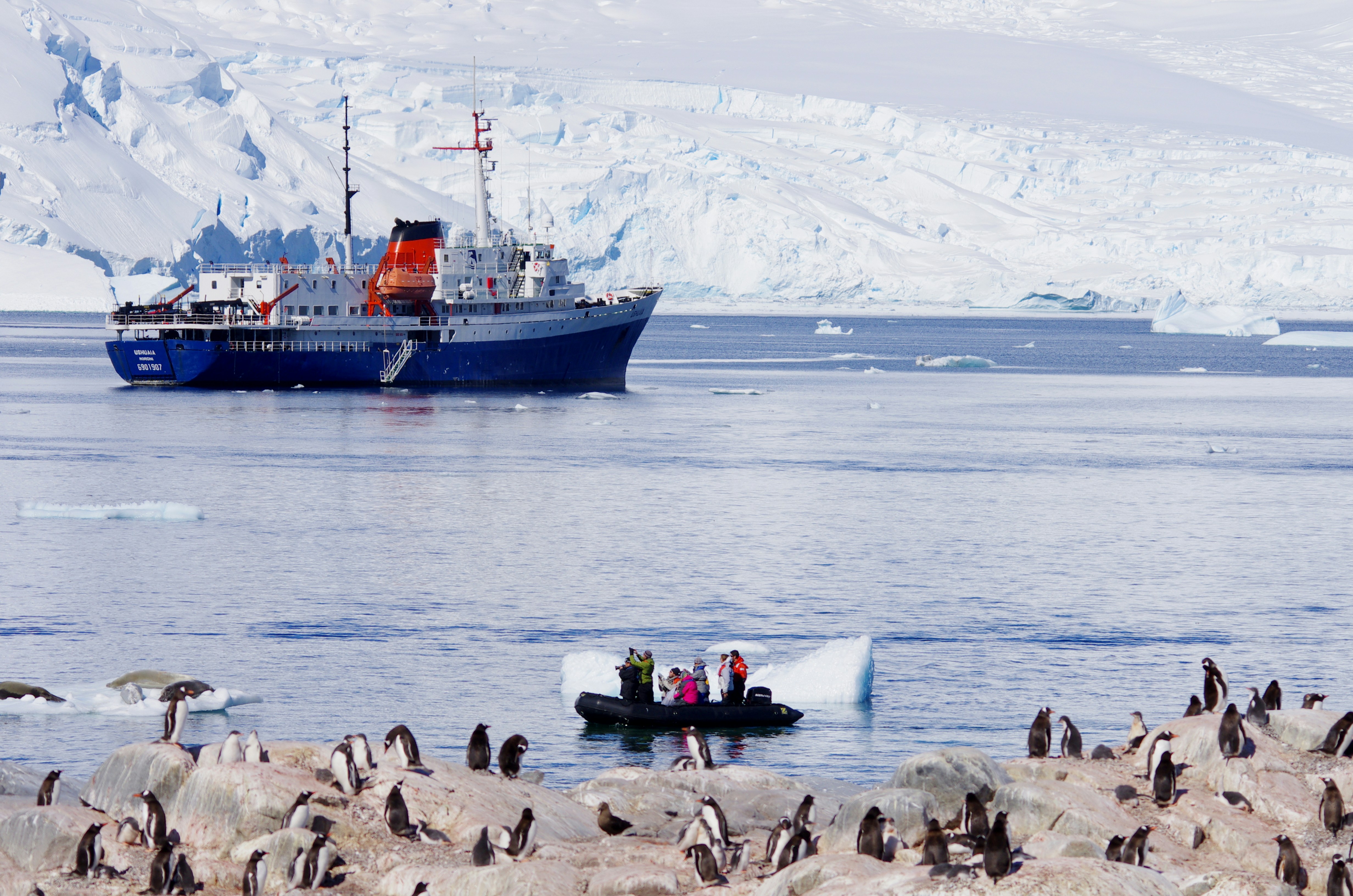 A blue research ship floats in icy Antarctic waters, with people in a small boat nearby. Dozens of penguins nest on the rocky shore, framed by snow-covered mountains—an incredible scene reminiscent of Svalbard vs. Greenland vs. Canadian Arctic vistas.