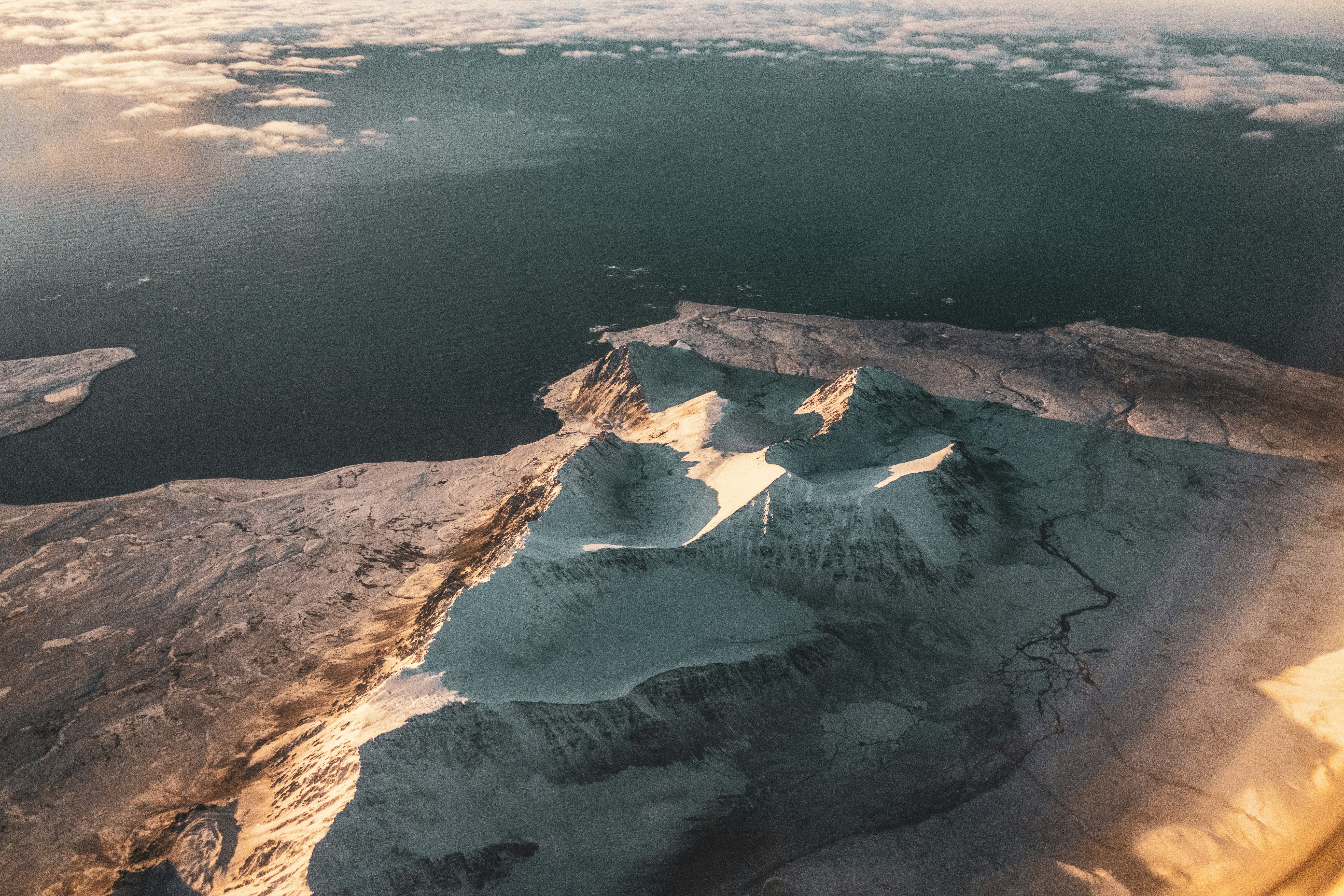 Aerial view of snow-covered mountains with sharp peaks and ridges beside a calm, dark sea under a cloudy sky, illuminated by warm, golden sunlight—home to unique species of penguins in Antarctica.