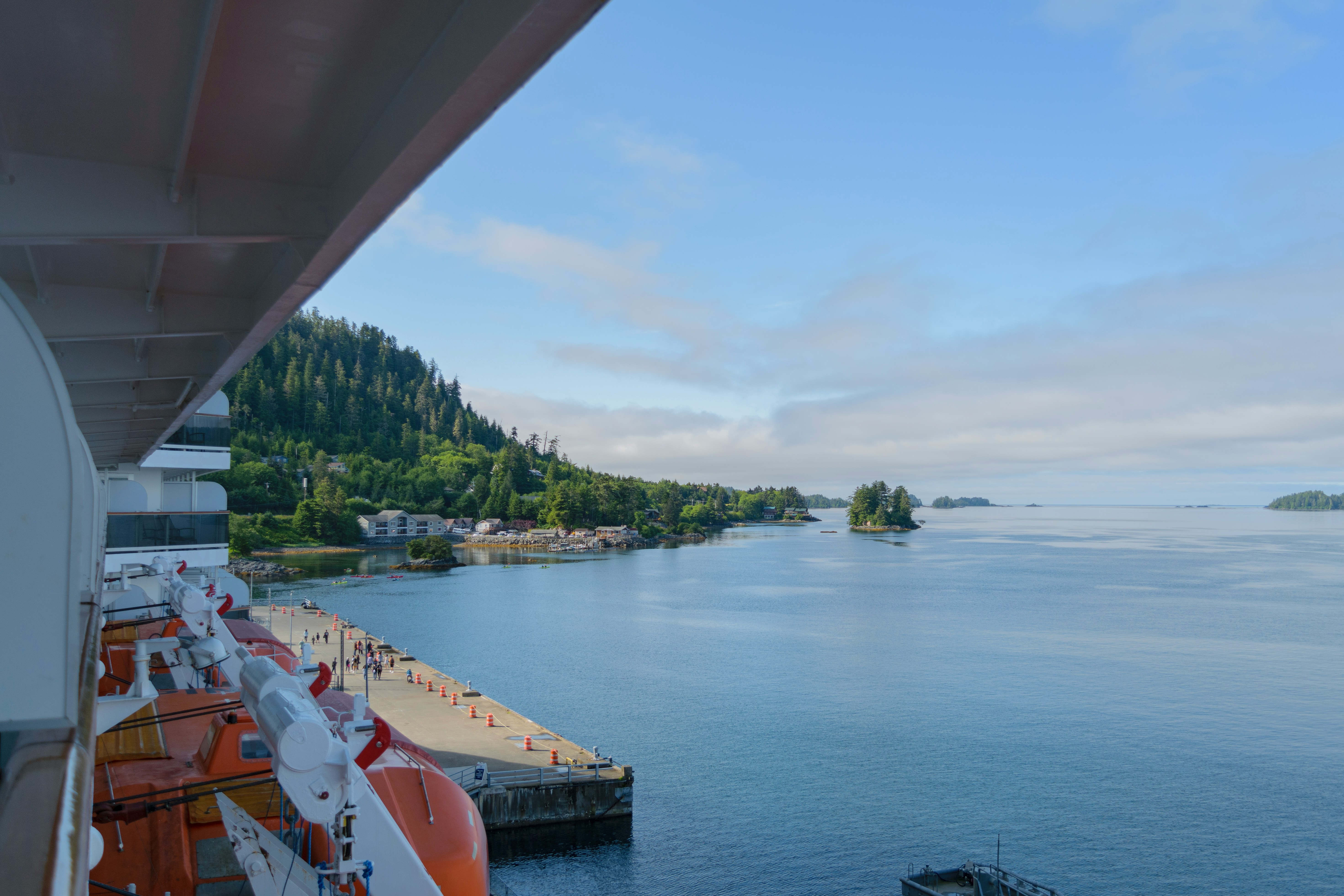 View from a cruise ship docked at a calm port—orange lifeboats in sight. The dock leads to a small settlement, framed by forested hills and blue water beneath a partly cloudy sky. Experience the Best Alaska Cruise 2025 adventure here.