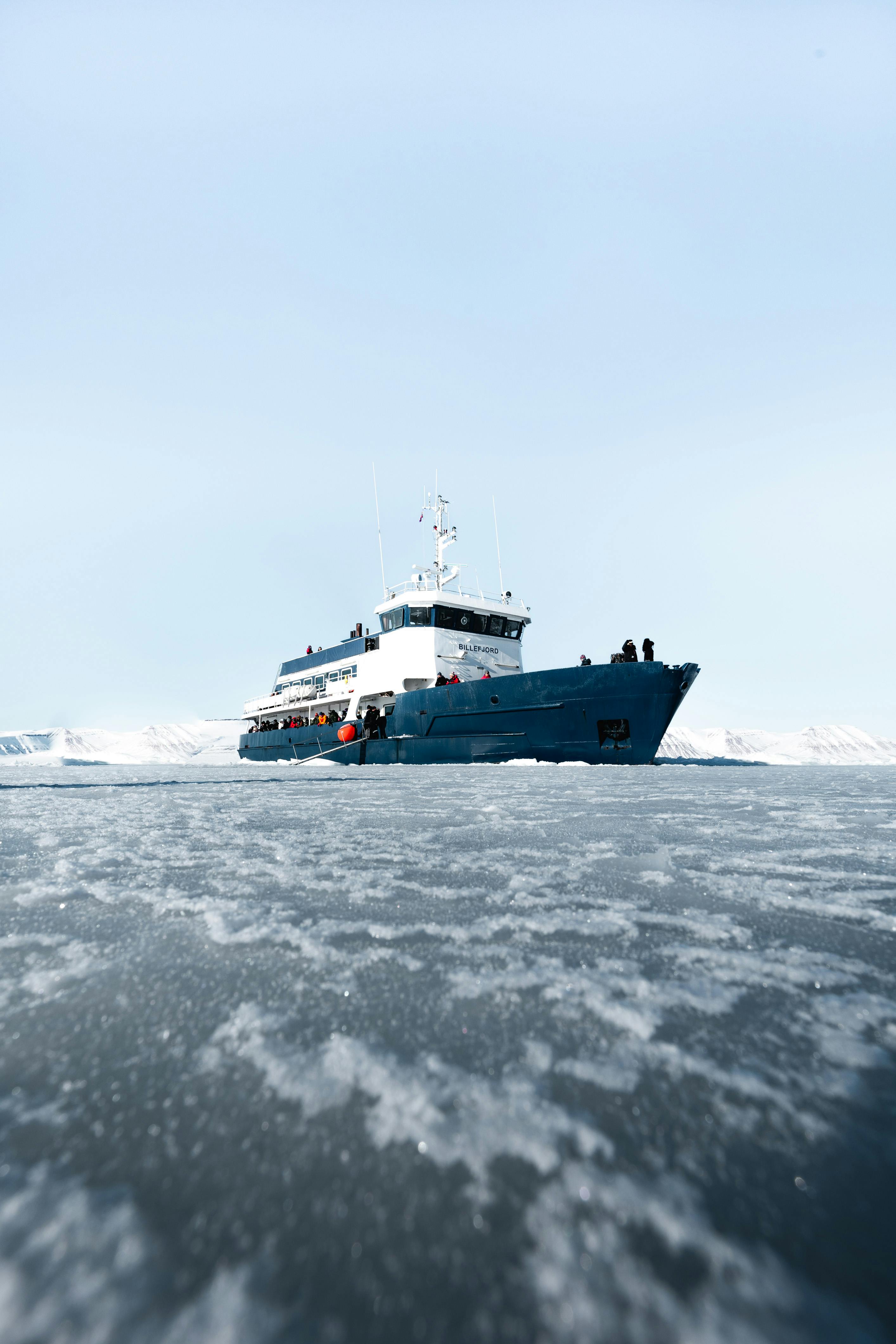 A blue ship navigates through icy waters, surrounded by snow-covered land under a clear sky. People are visible on the deck, enjoying an unforgettable journey typical of Luxury Small Ship Alaska Cruises in this polar region.