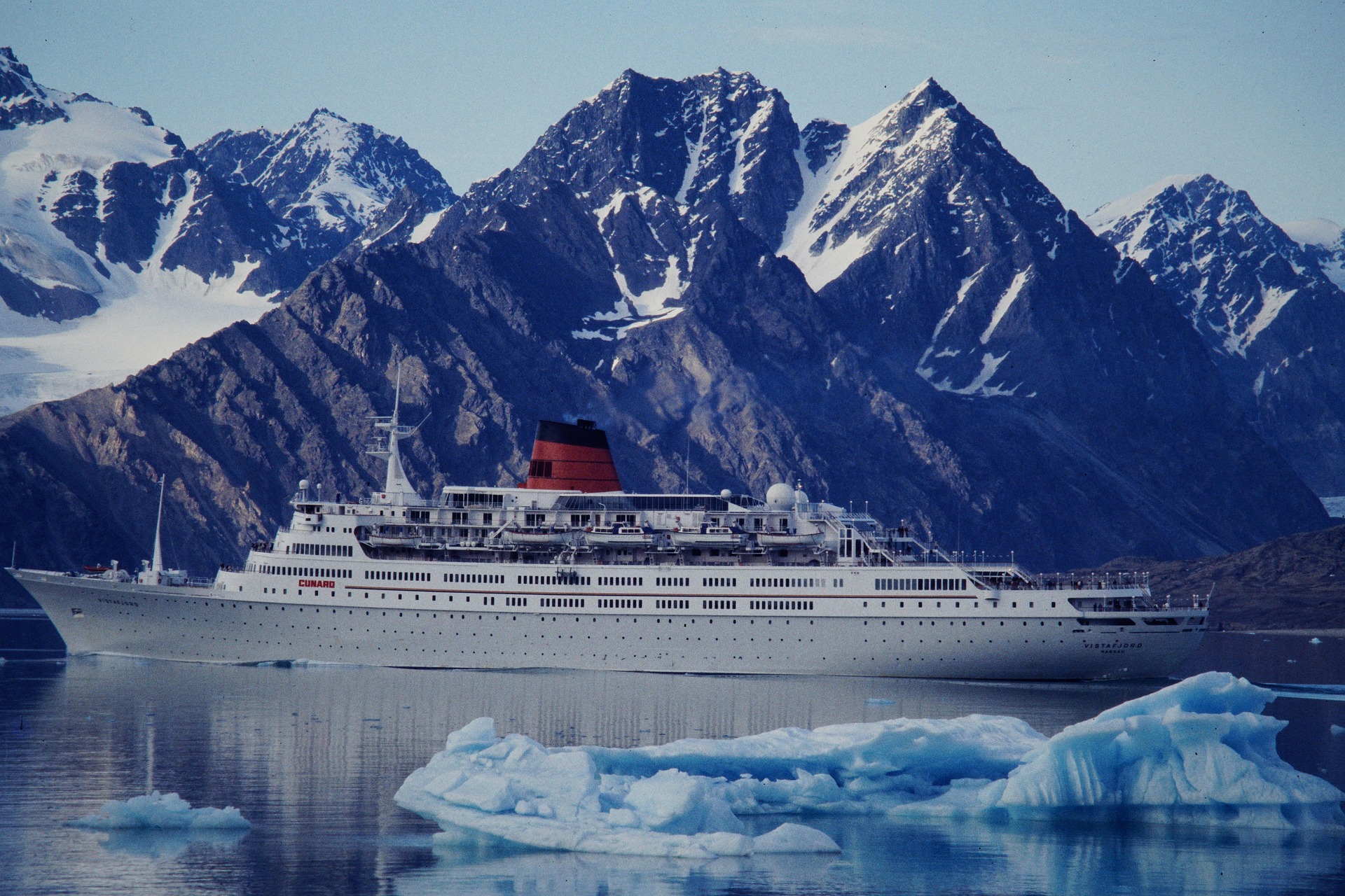 A large white cruise ship sails through calm icy waters with floating icebergs, set against rugged snow-capped mountains—a classic scene when exploring the Svalbard vs. Greenland vs. Canadian Arctic debate among travelers.