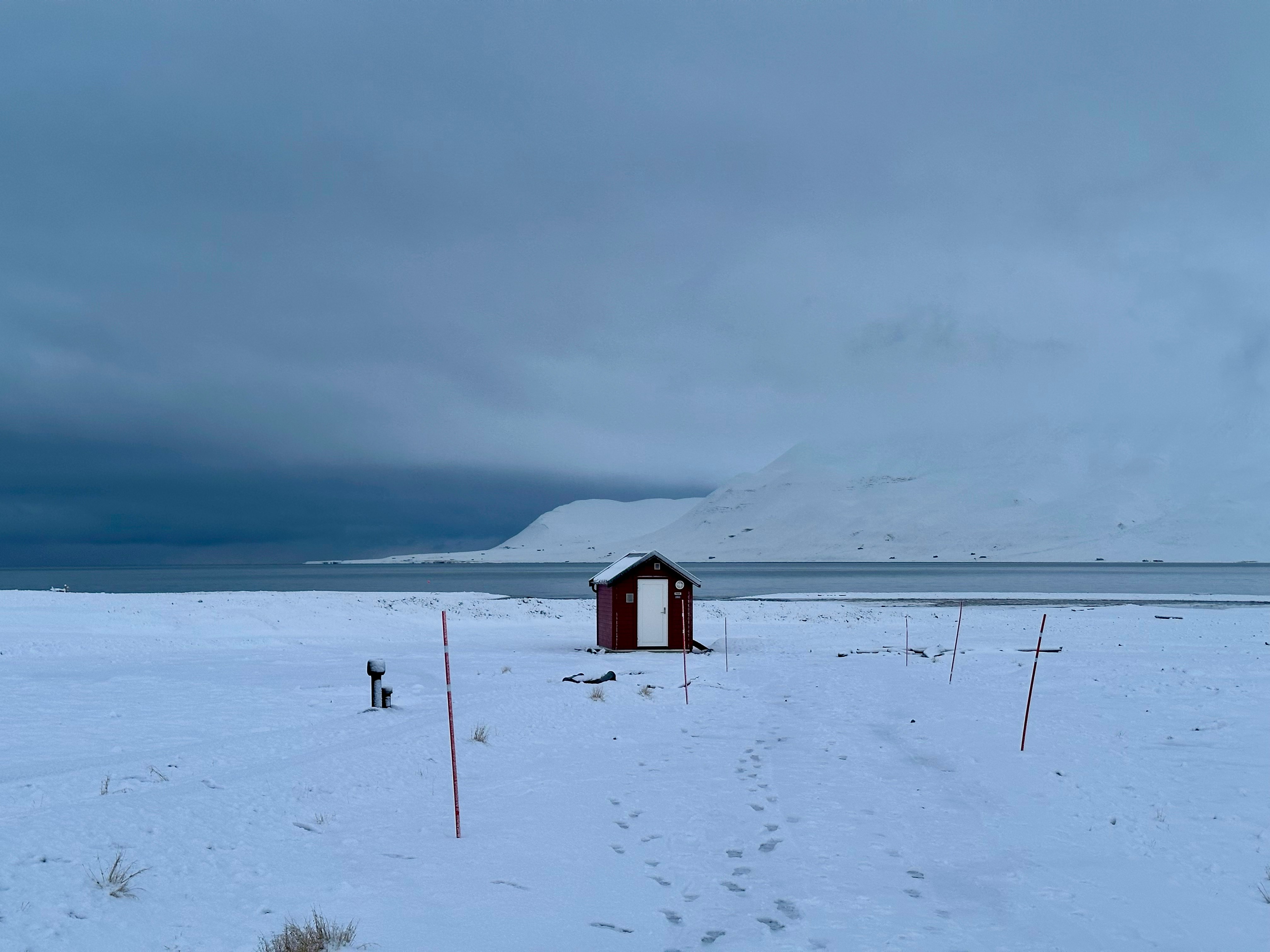 A small red cabin stands alone on a snowy landscape by the water, with snow-covered mountains in the background under a cloudy sky—evoking the remote beauty found in Svalbard vs. Greenland vs. Canadian Arctic wilderness. Faint tracks lead to the cabin.