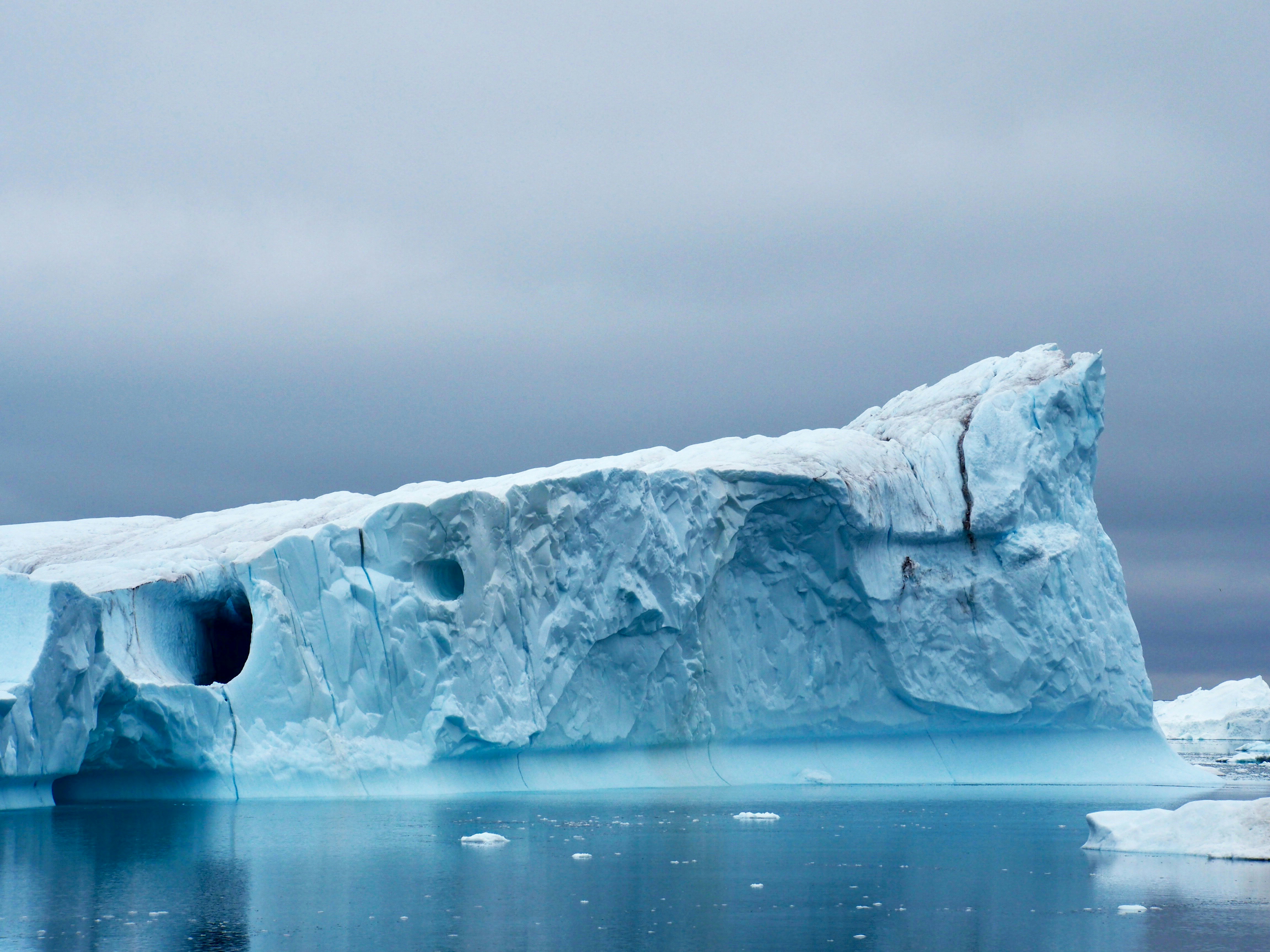 A large iceberg with jagged edges floats in calm, icy water beneath a cloudy, gray sky—a scene reminiscent of Svalbard vs. Greenland vs. Canadian Arctic landscapes, with a dark hollow, cracks, and scattered ice pieces on the water’s surface.