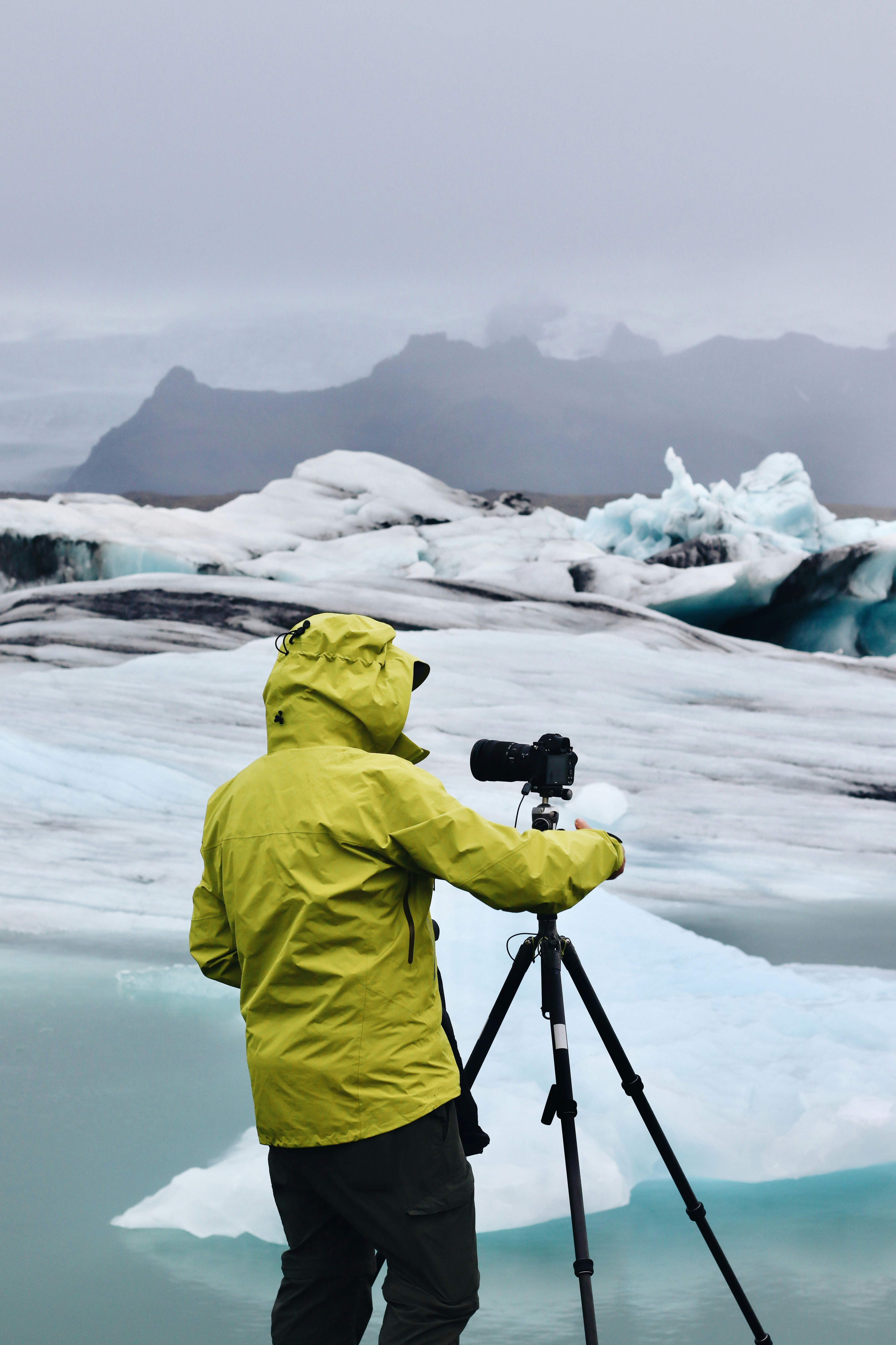 A person in a yellow jacket stands by icy water, camera on tripod, capturing glaciers and snow-covered mountains—scenery often wondered about by those asking, How Much Does an Alaska Cruise Cost?—beneath a cloudy sky.