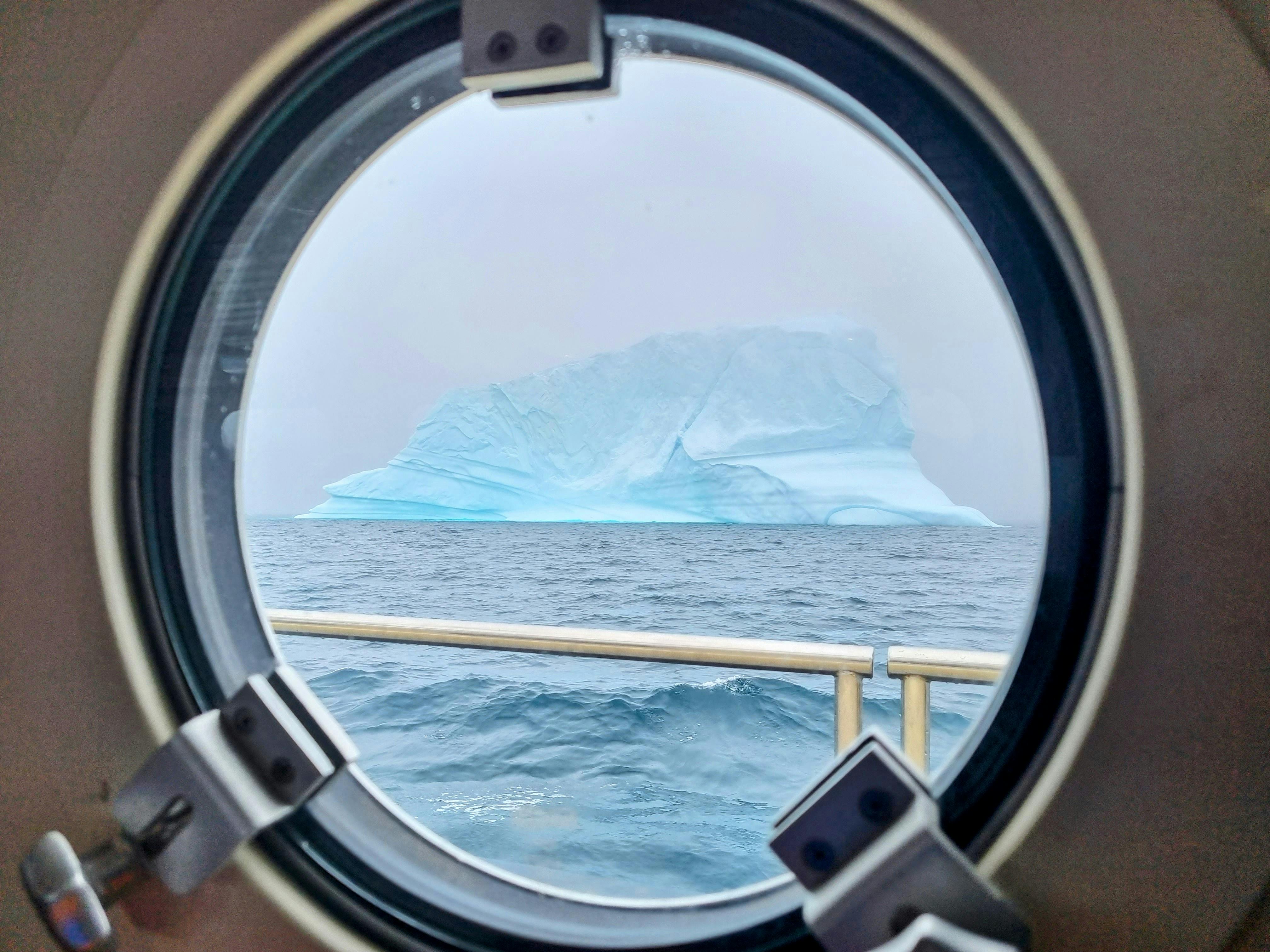 A large iceberg floats on the ocean, seen through a round porthole window from inside a ship—perfect scenery for an Antarctica Cruise for Wildlife Photographers, with cloudy skies and part of the ship’s railing visible.