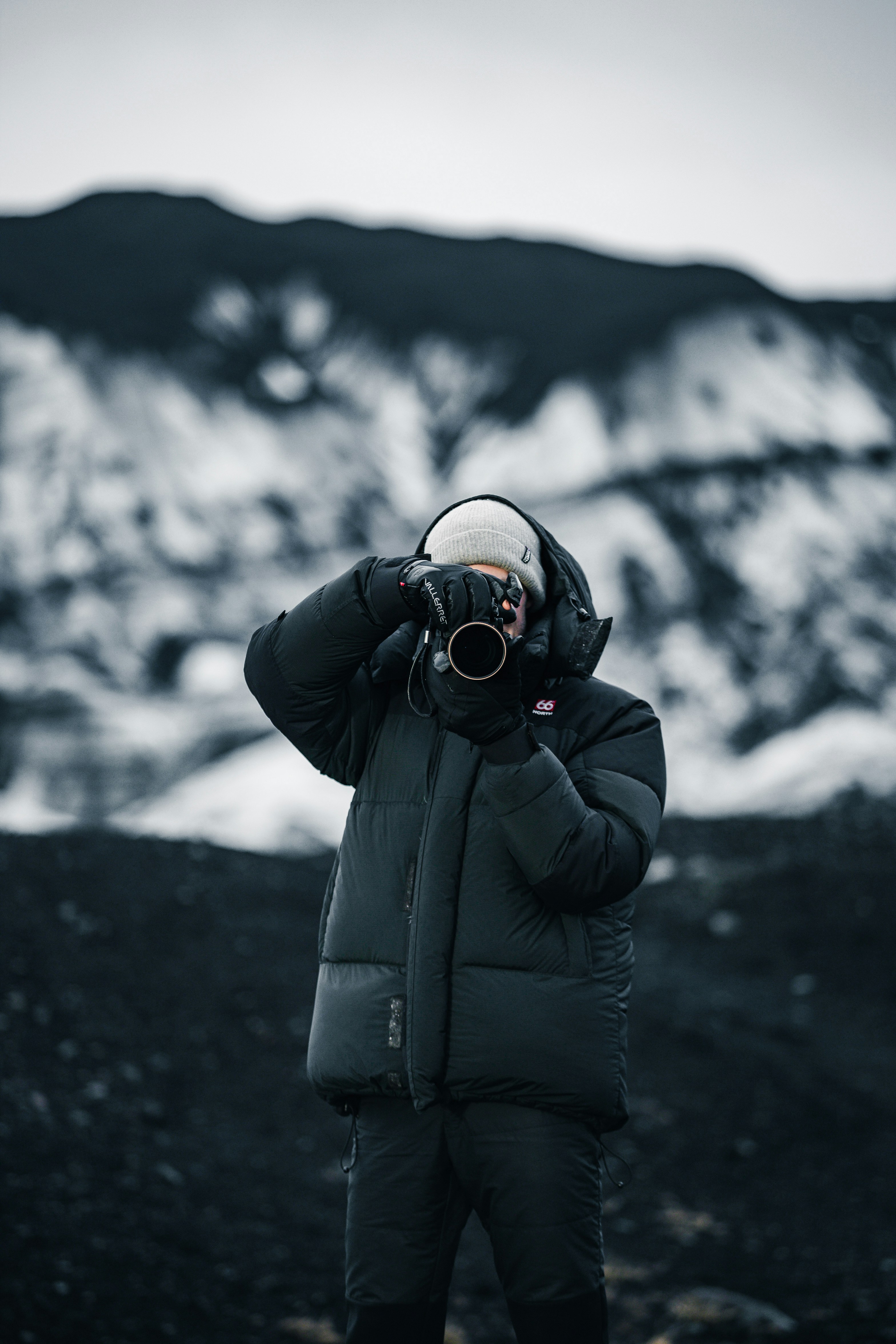 A person in a dark puffer jacket and light beanie takes a photo with a camera outdoors amid snow-dusted mountains, capturing the spirit of adventure that makes many wonder, how much does an Alaska cruise cost?.