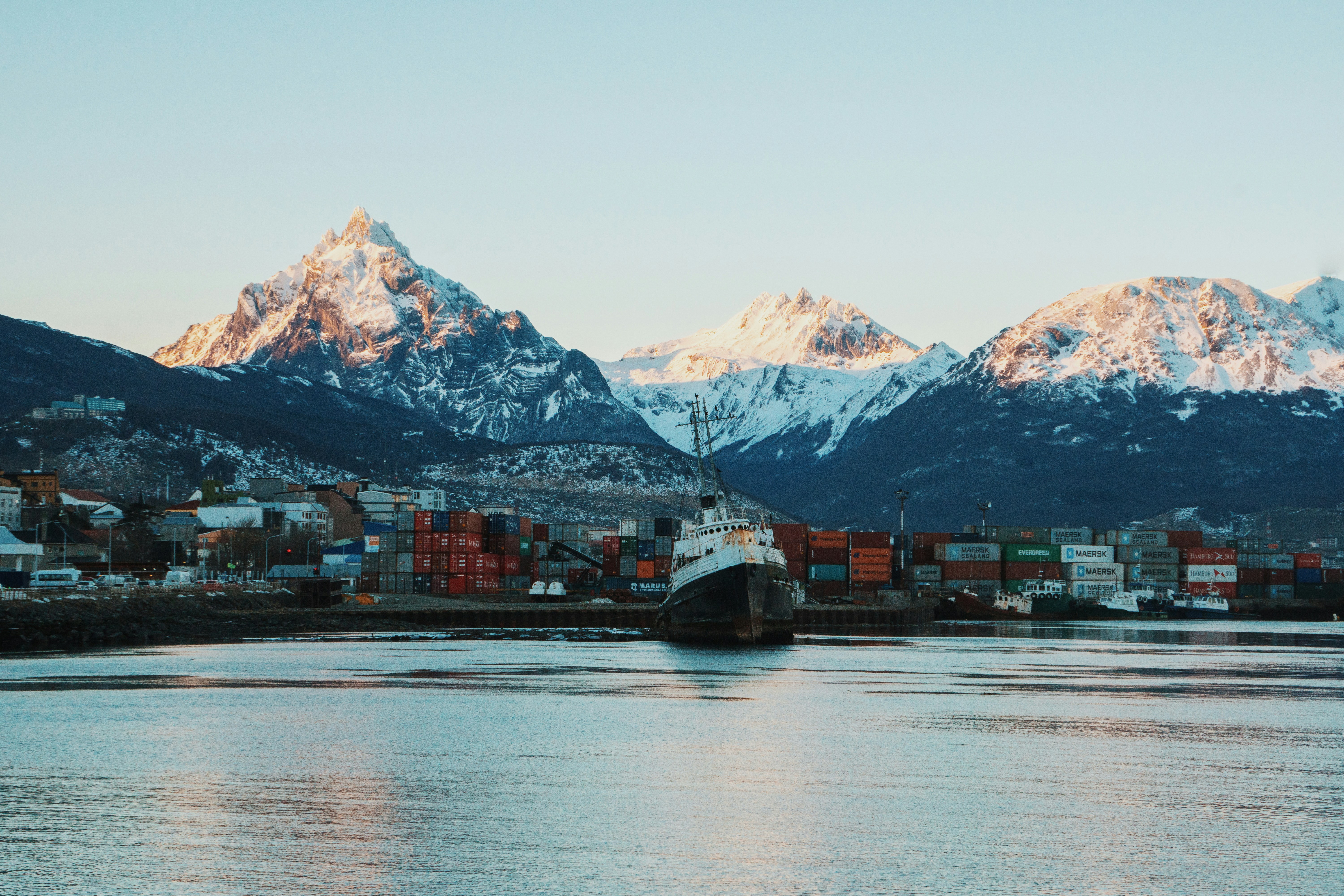 A cargo ship is docked at a port with colorful shipping containers, set against snow-capped mountains and calm waters—typical scenery when exploring Svalbard vs. Greenland vs. Canadian Arctic regions.