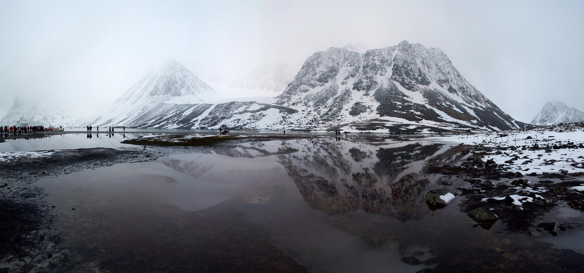 Snow-covered mountains reflected in a still, shallow lake under a cloudy sky evoke the raw beauty found in Magdalen Bay in Spitsbergen, as small groups along the shoreline highlight the vast scale of this rugged, wintry landscape.