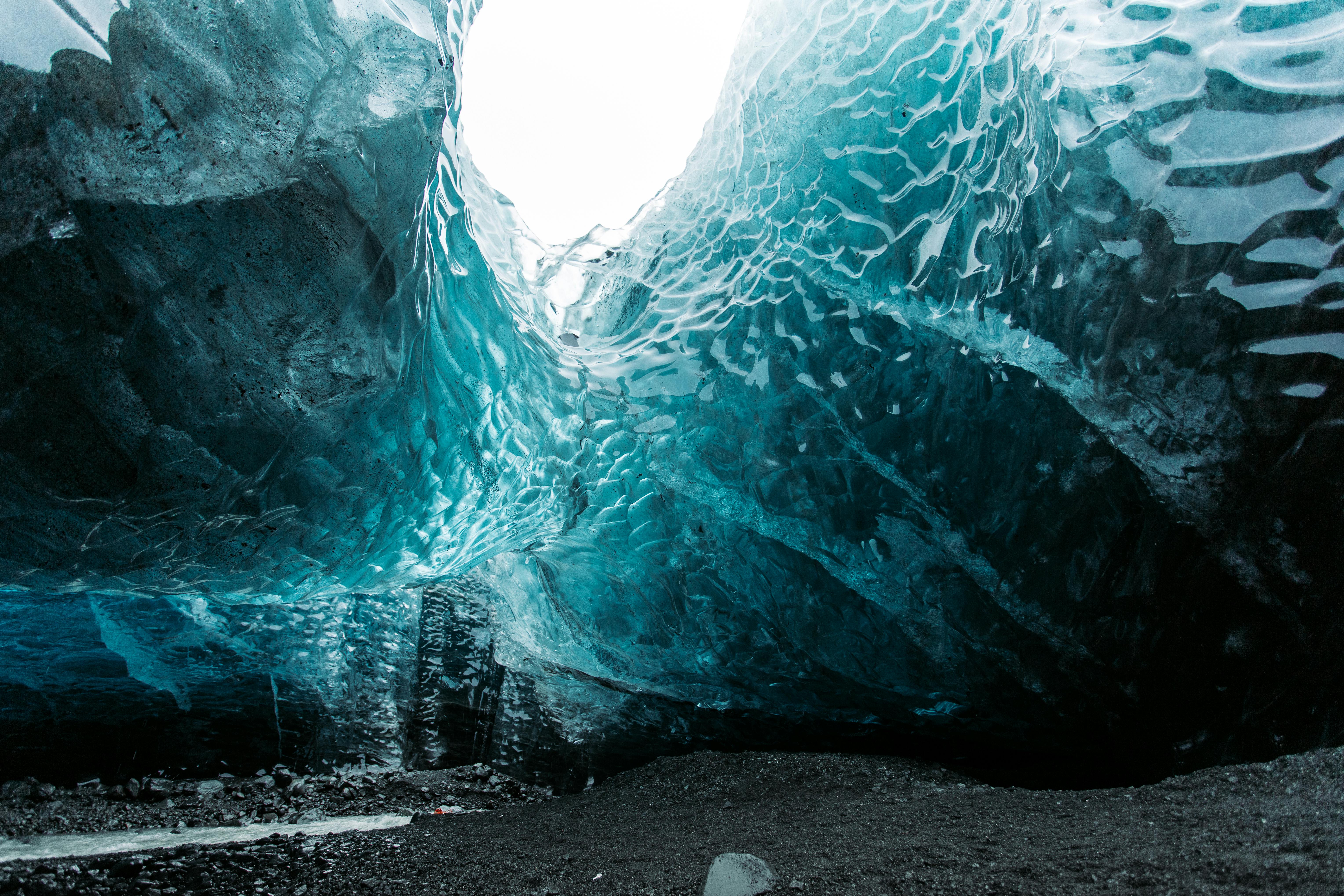 View from inside an ice cave, with smooth, blue-tinted ice walls and a bright opening above—a breathtaking scene often admired on the Best Alaska Cruises for 2026—contrasting against the dark, rocky ground below.