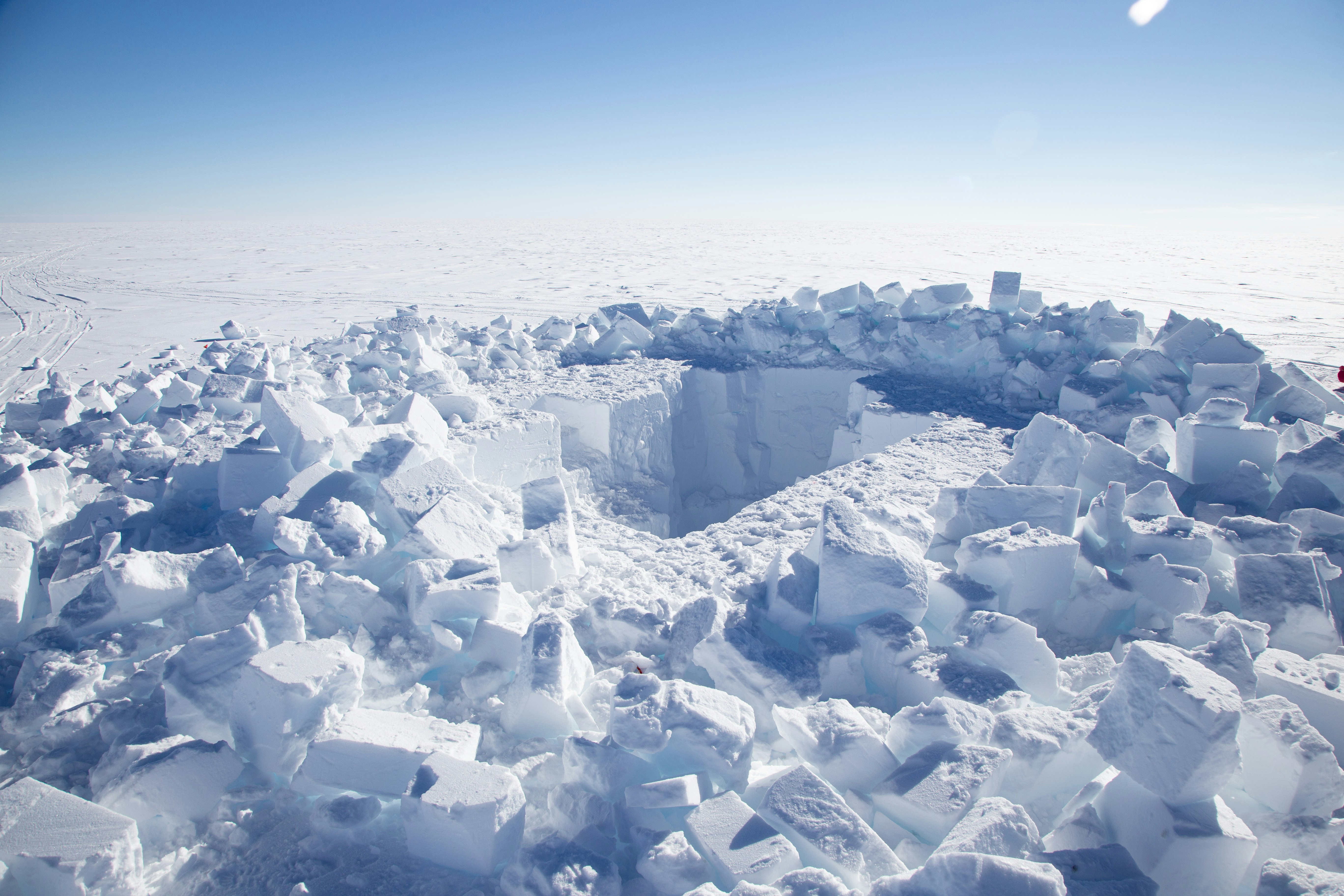 A large rectangular hole is cut into a snowy, frozen landscape. Neatly stacked blocks of snow surround the hole, with an endless expanse of snow and a clear blue sky in the background.
