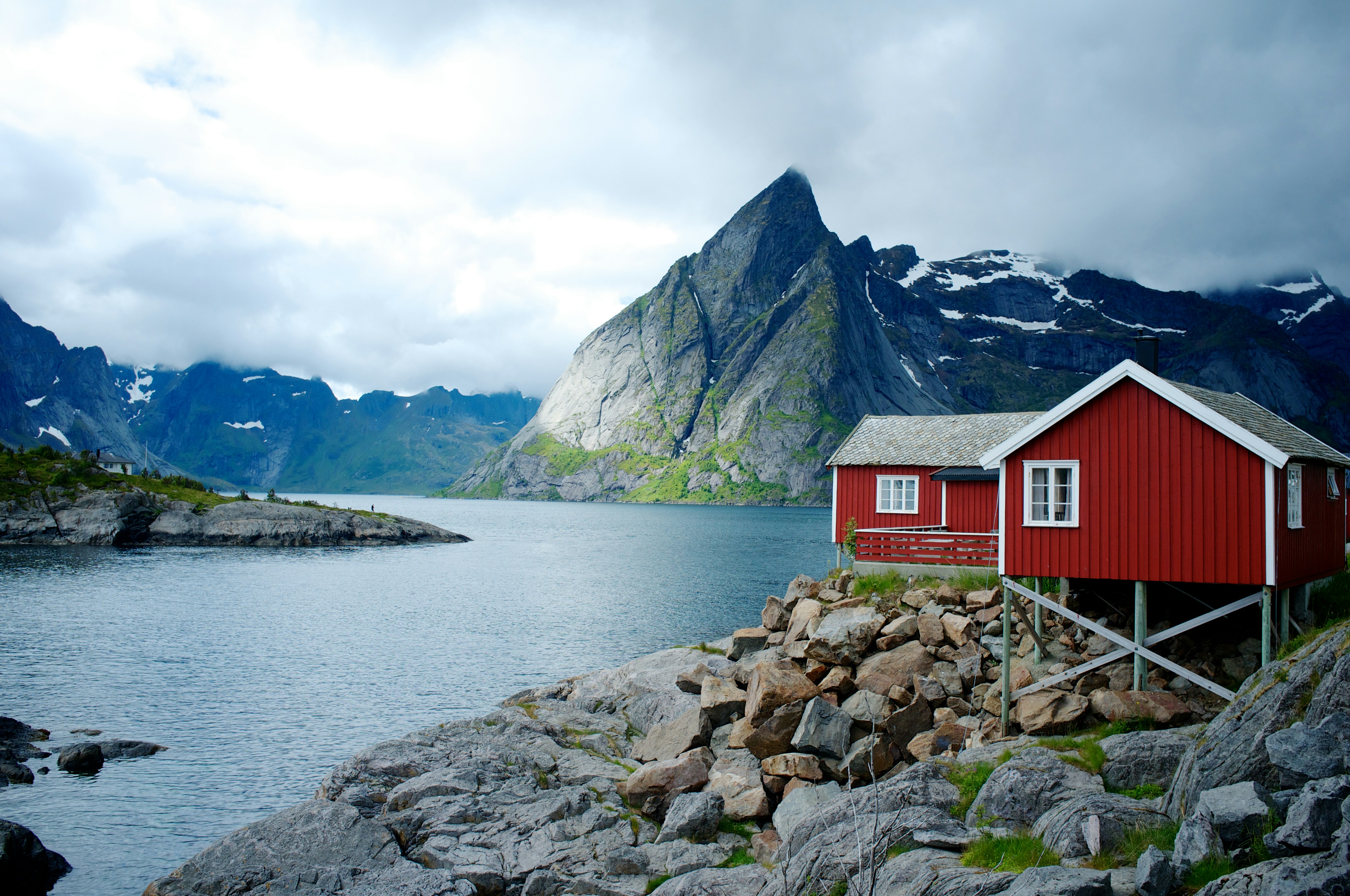 A red wooden cabin on a rocky shore overlooks a calm fjord, surrounded by mountains under a cloudy sky—reminiscent of the scenery seen on Small Ship Cruises to Alaska, with snow patches visible on distant peaks.